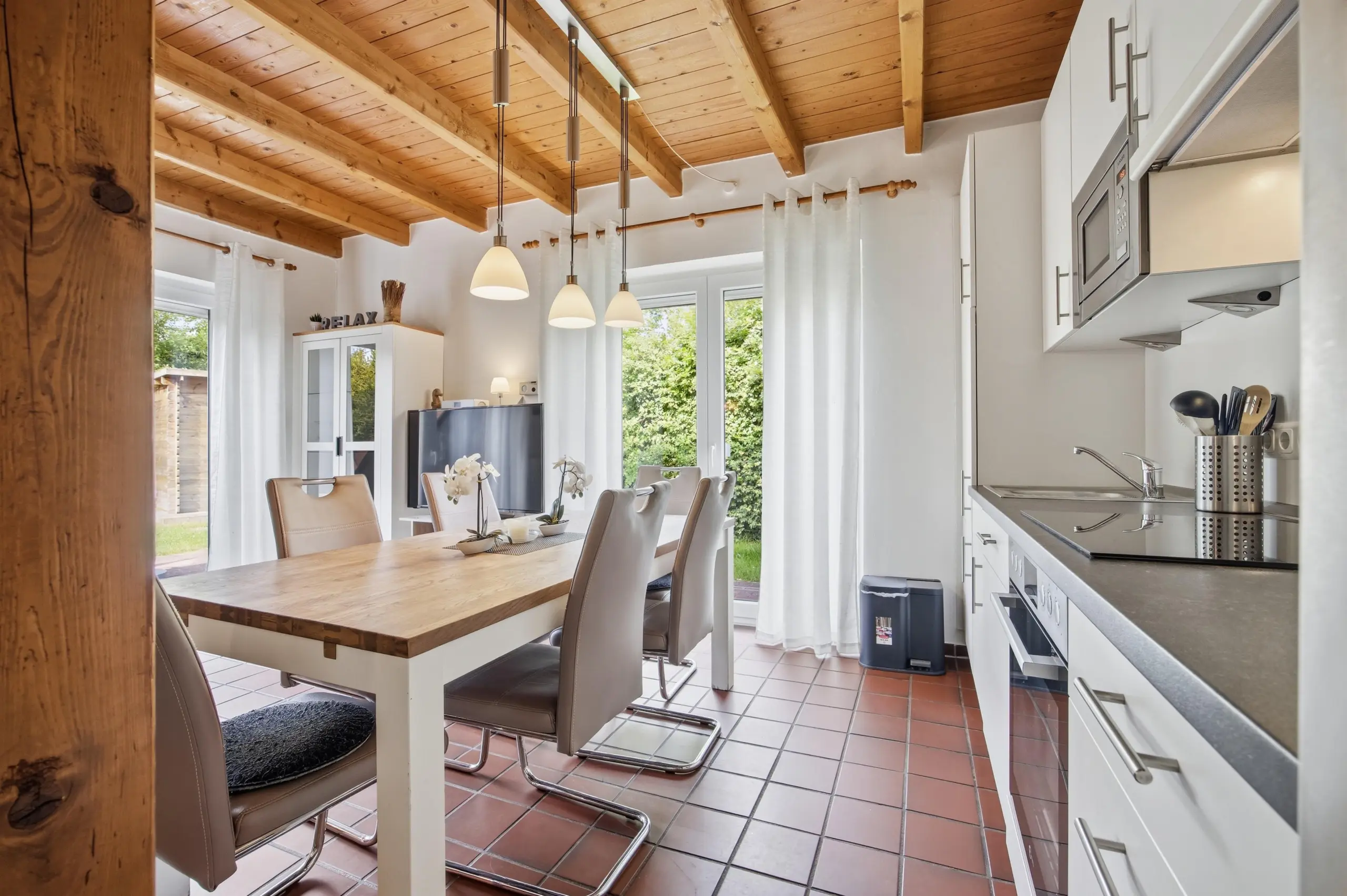 Open-plan kitchen and dining area with exposed wooden ceiling beams, pendant lights over a wooden table, and white cabinets along one wall.