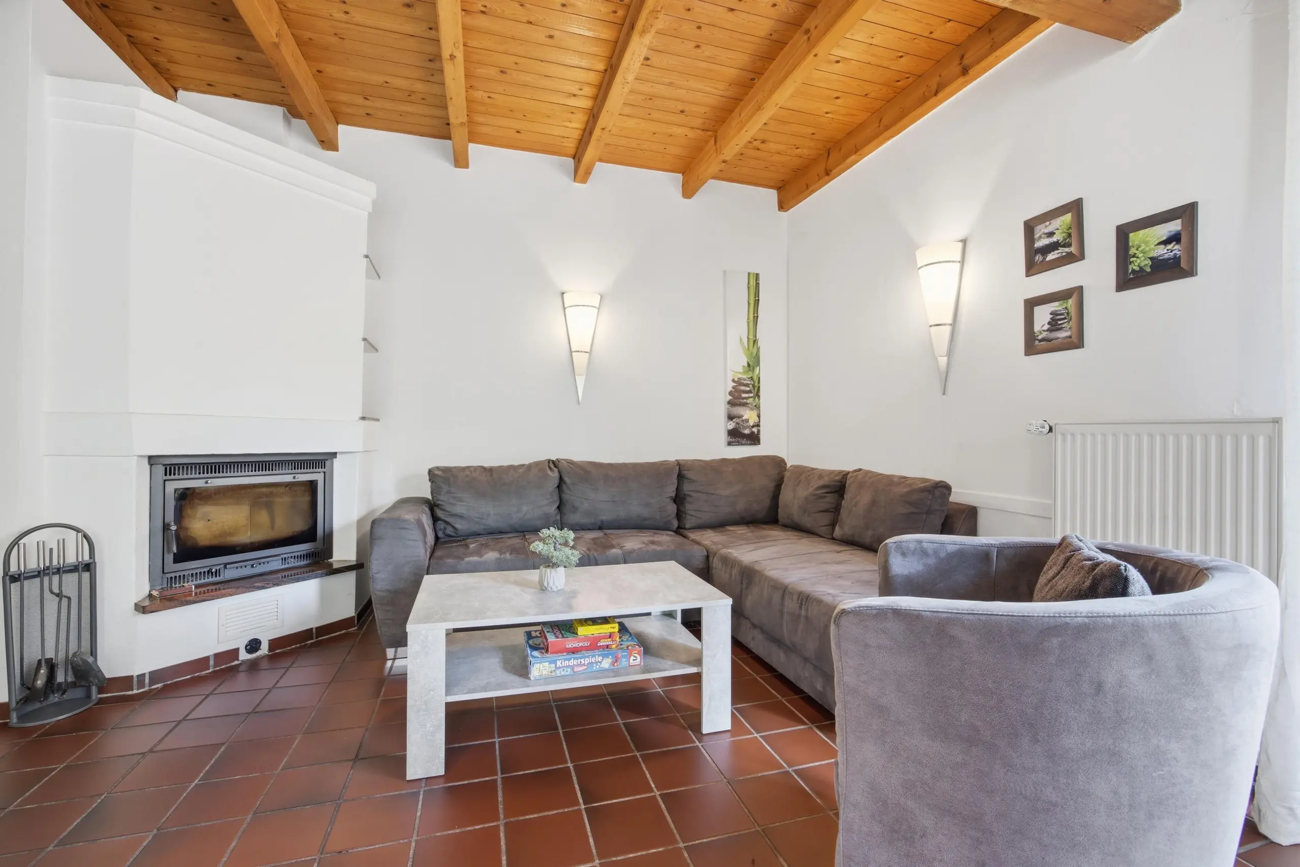 Living room with brown tiled floor, white walls, wooden beam ceiling, gray sectional sofa, white fireplace on the left, and a coffee table with wall art.
