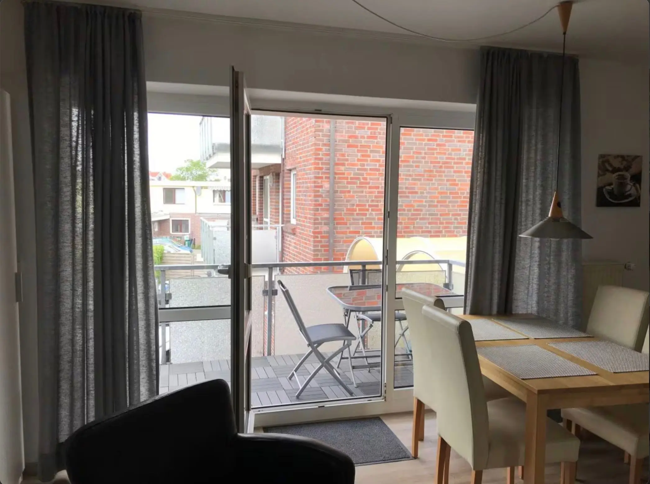 Open glass balcony door with metal chairs and a table outside, seen from a living room with dark curtains