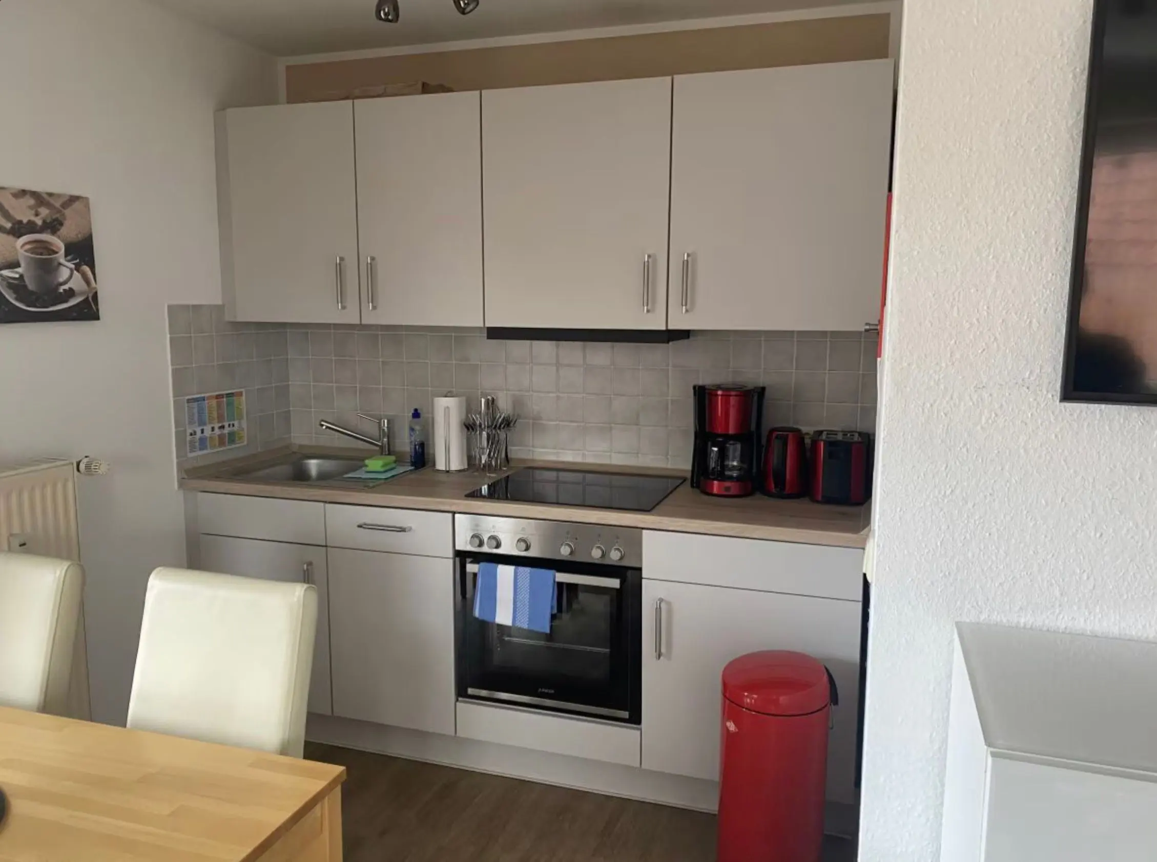 Modern kitchen with grey upper cabinets, stainless sink, electric stove, and red coffee machine and trash bin on the counter.