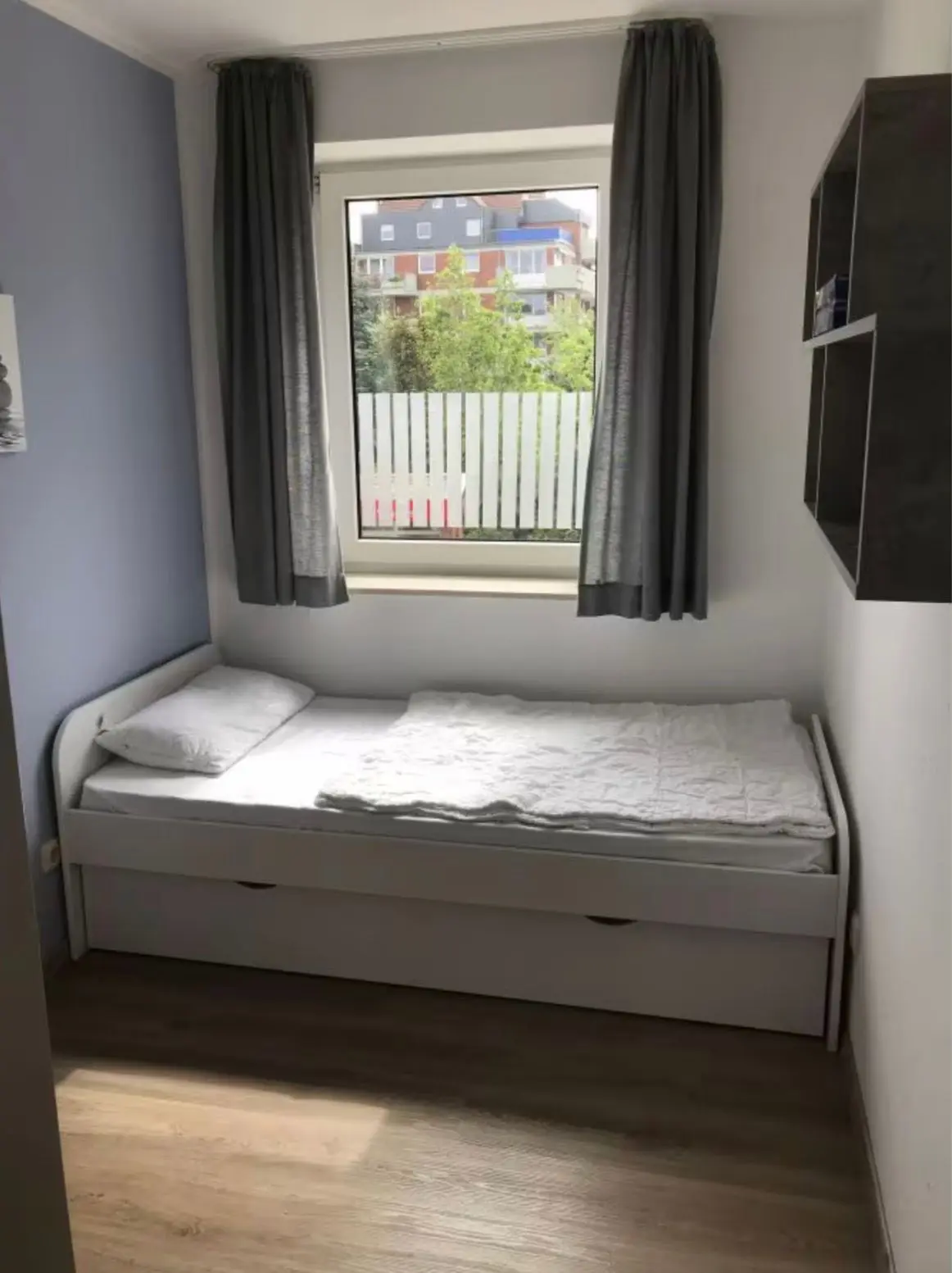 Small bedroom with a white single bed under a window, gray curtains, and a wall shelf on the right.