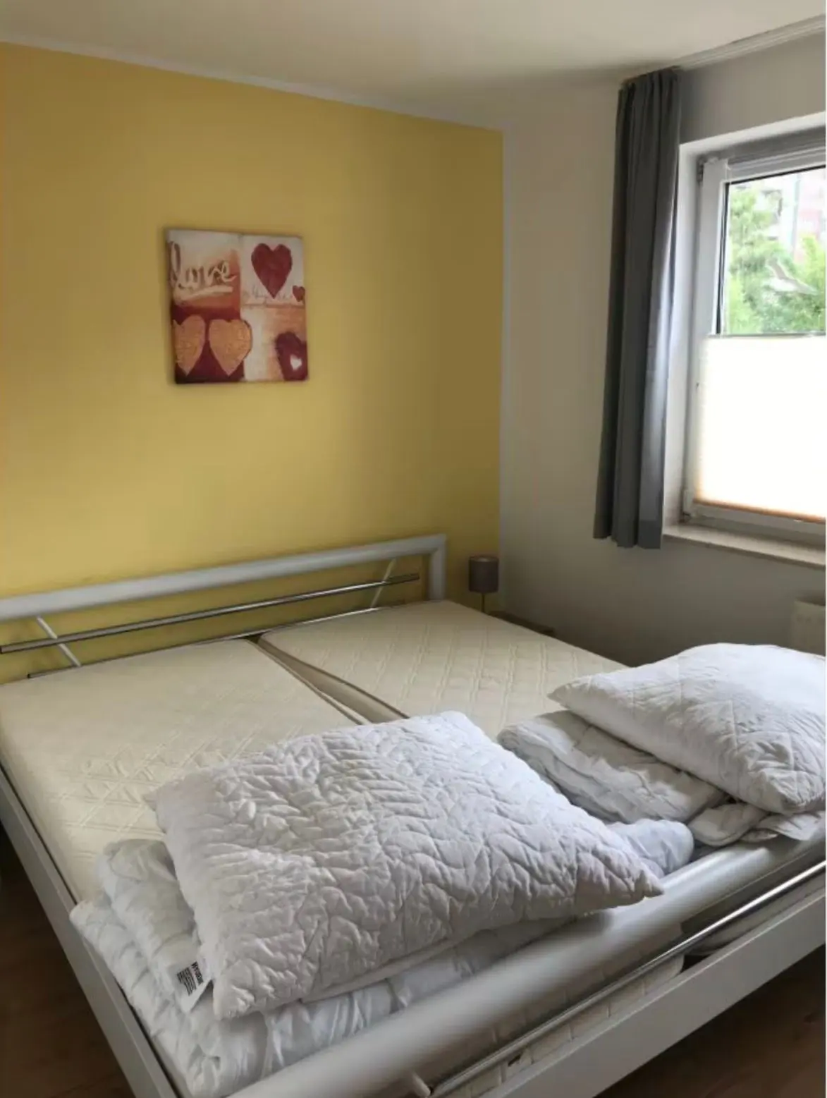 Bright yellow feature wall in a bedroom with a metal-framed bed, white quilts stacked on top, and a window with gray curtains to the right.