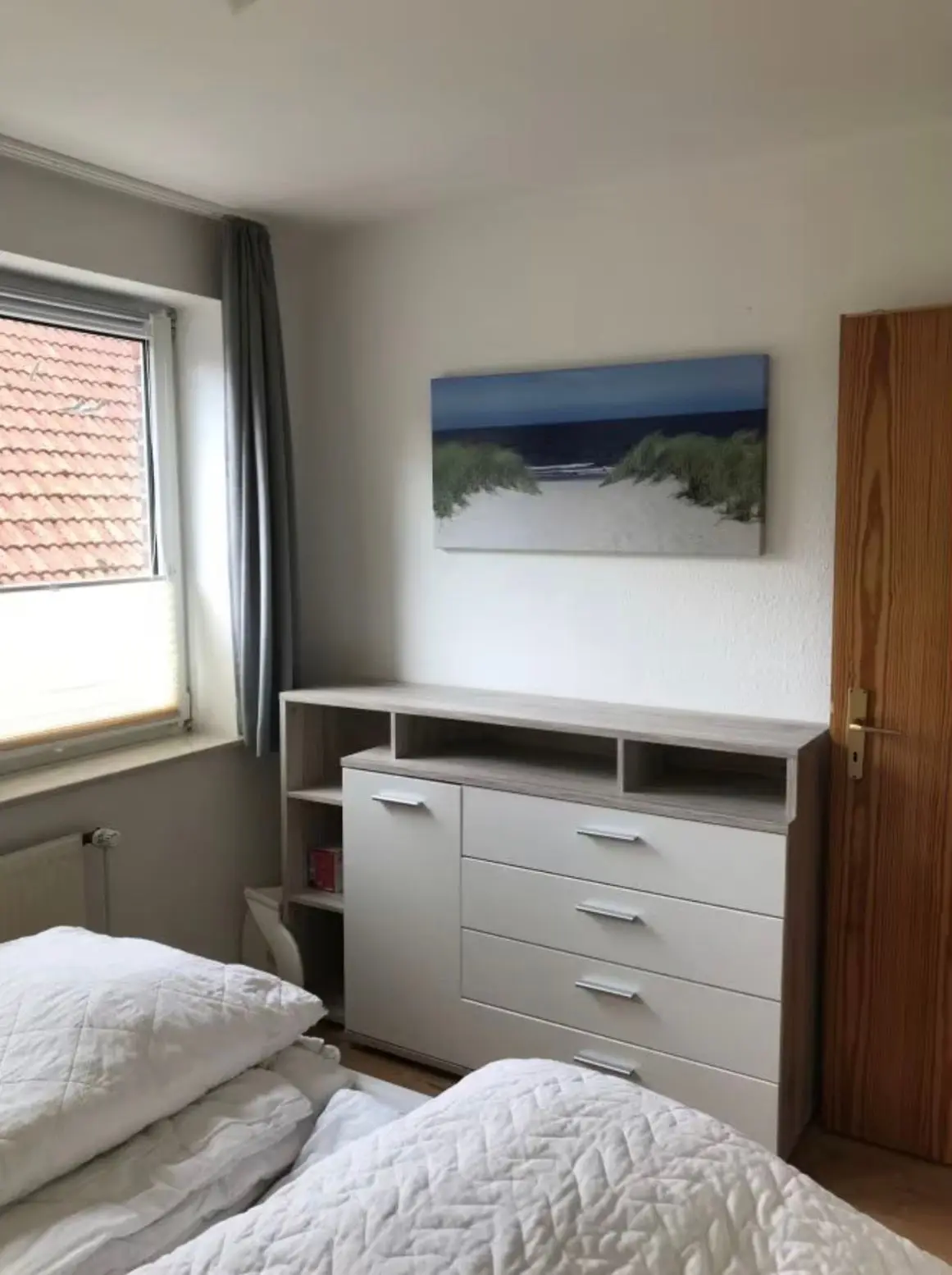 A bedroom corner with a light wood dresser featuring open shelves and white drawers, two beds with white quilts in the foreground, and a beach photo on the wall above.