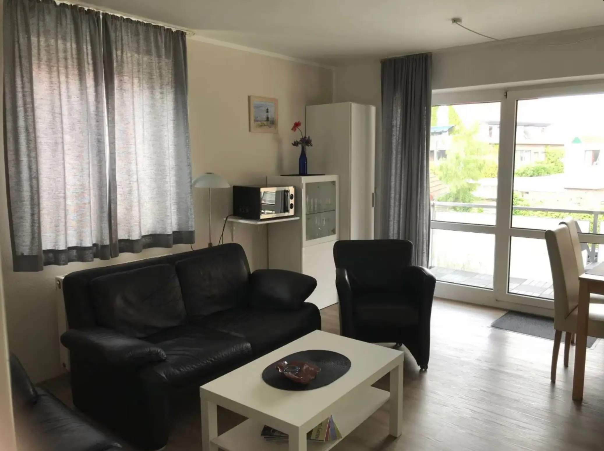 Living room with black leather sofa, white coffee table, and a balcony door with silver curtains seen through large glass windows.
