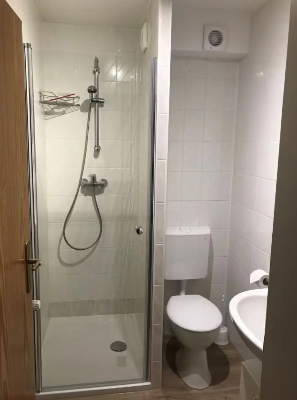 Compact bathroom with glass shower enclosure on the left, toilet and sink on the right against white tiled walls.