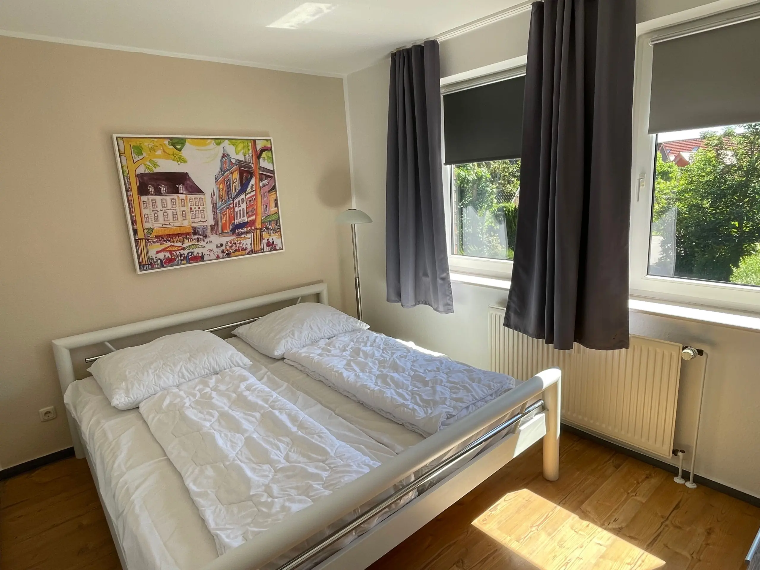Twin beds with white bedding in a sunlit bedroom, a colorful wall painting, and a large window with dark blinds and curtains.