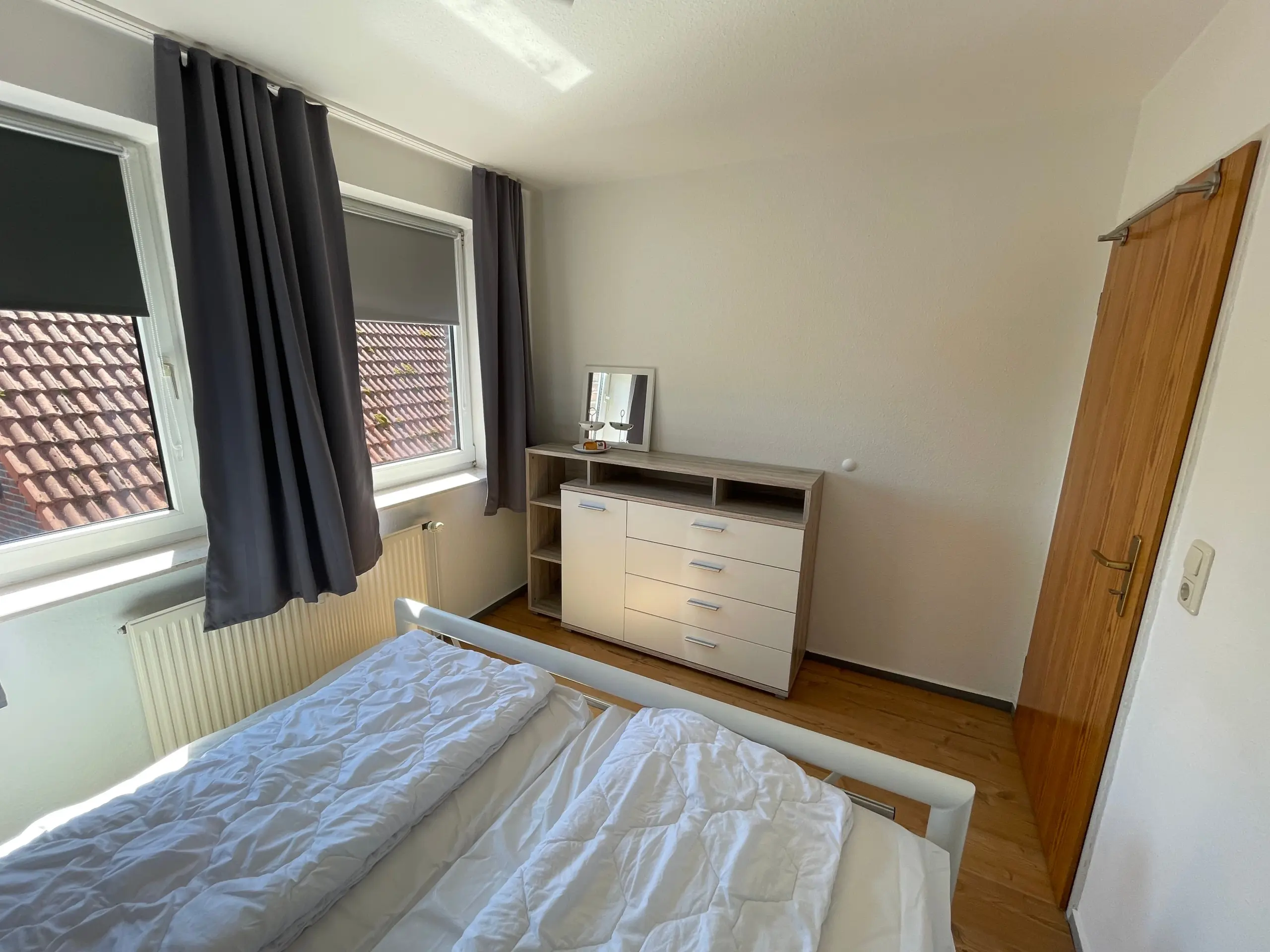 Bedroom corner with two single beds wearing white duvets, a wooden dresser with a small mirror, and dark curtains by a window.