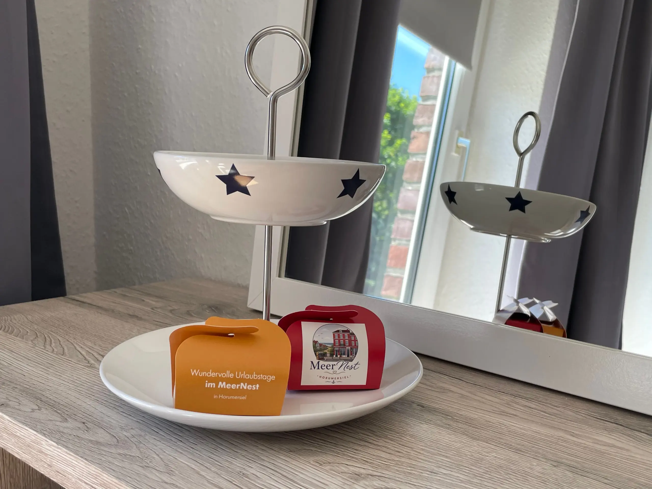 Two white bowls with blue stars on a metal stand, set on a wooden table by a window; orange and red promotional cards nearby for MeerNest vacation housing.