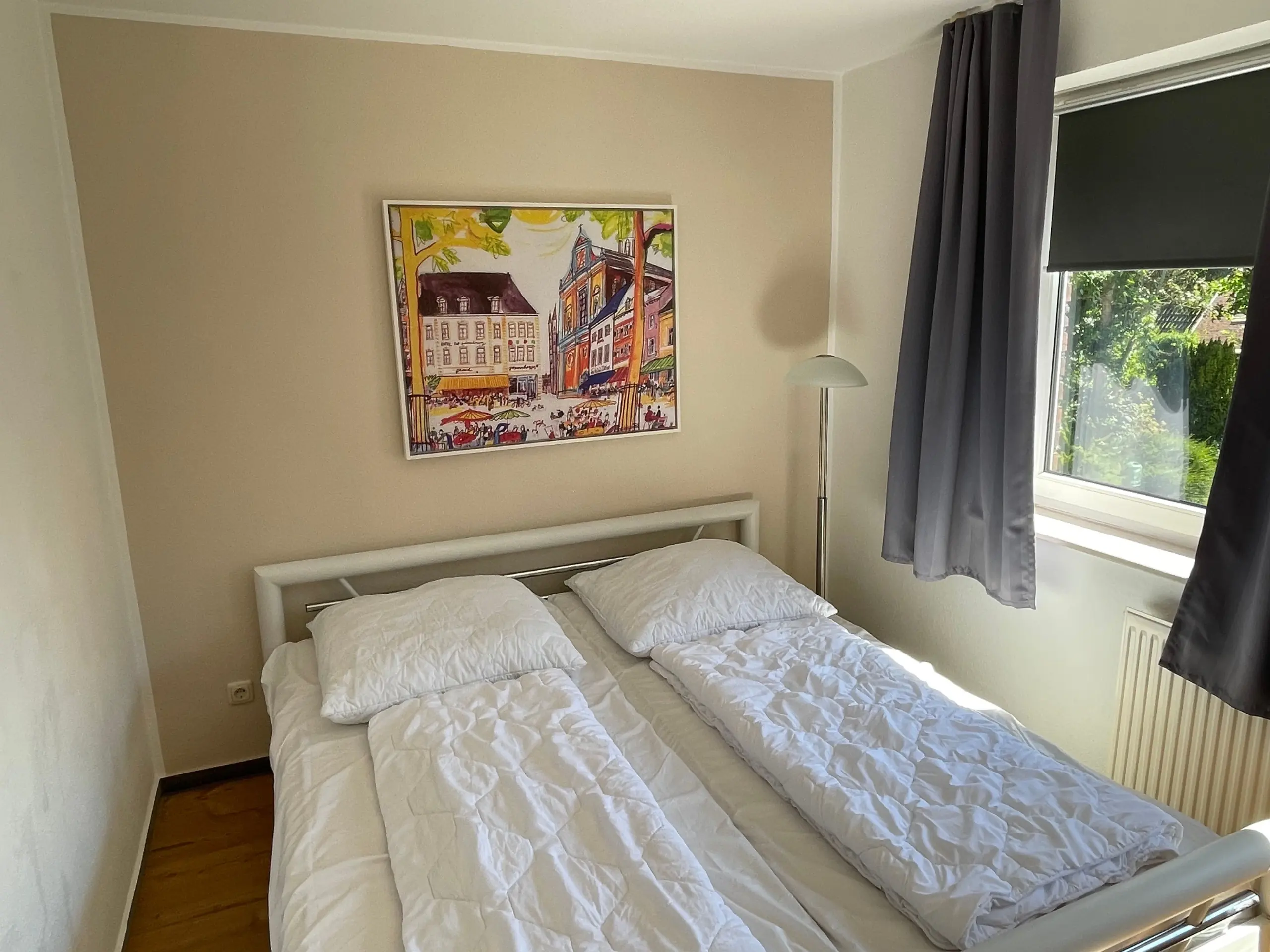 Twin bed in a light-colored bedroom with a framed city painting above and a standing lamp nearby, window with gray curtains to the right.