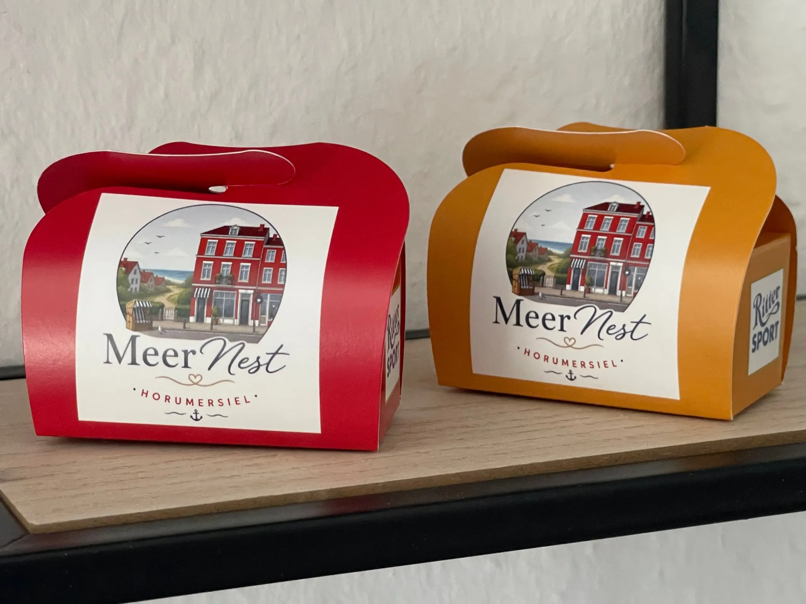 Two colorful gift bags on a wooden shelf, red on the left and orange on the right, each with a white MeerNest label.