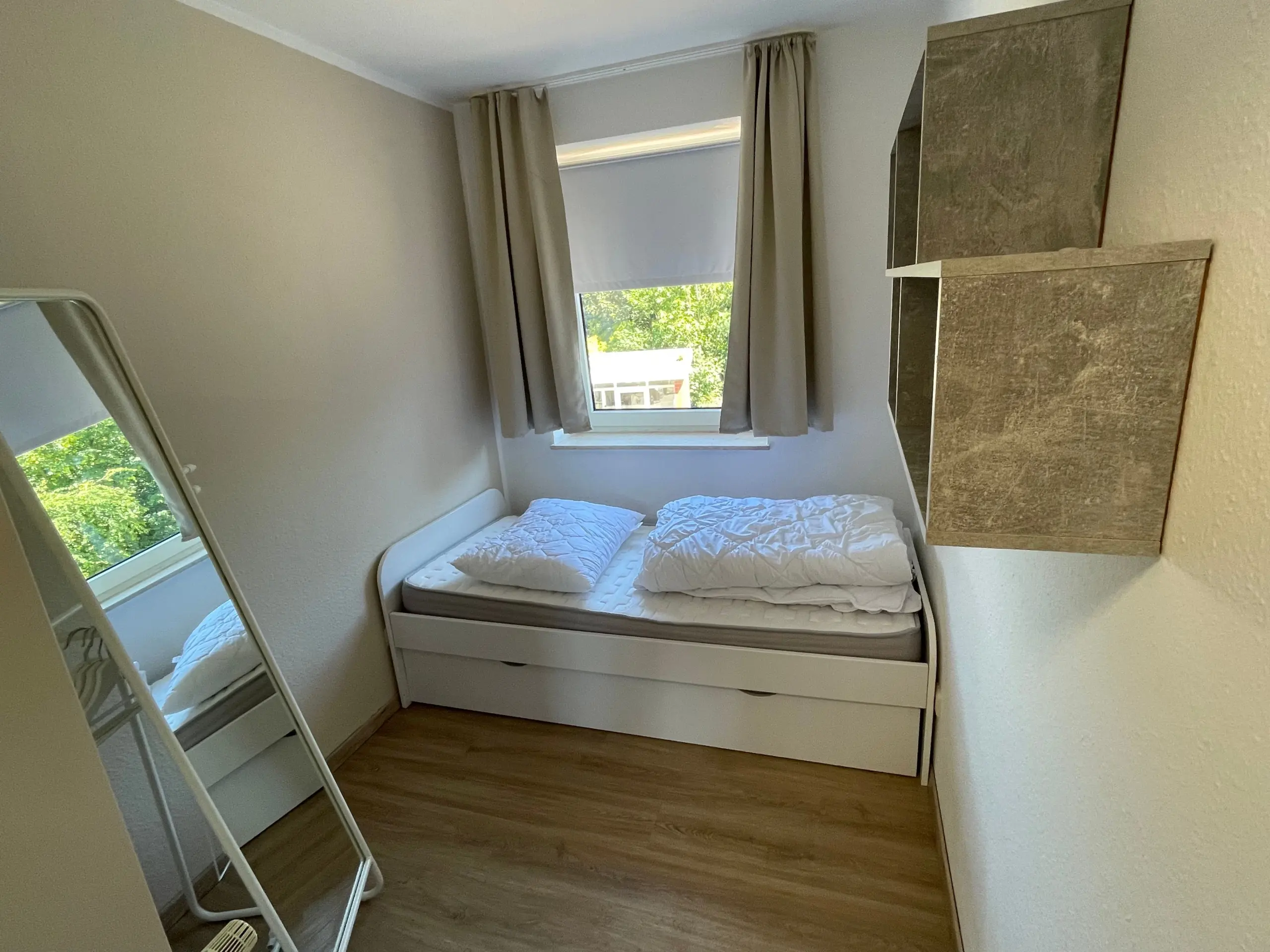 Small bedroom with a white bed, two pillows and folded quilt, window letting in greenery, beige walls, and light wood floor.