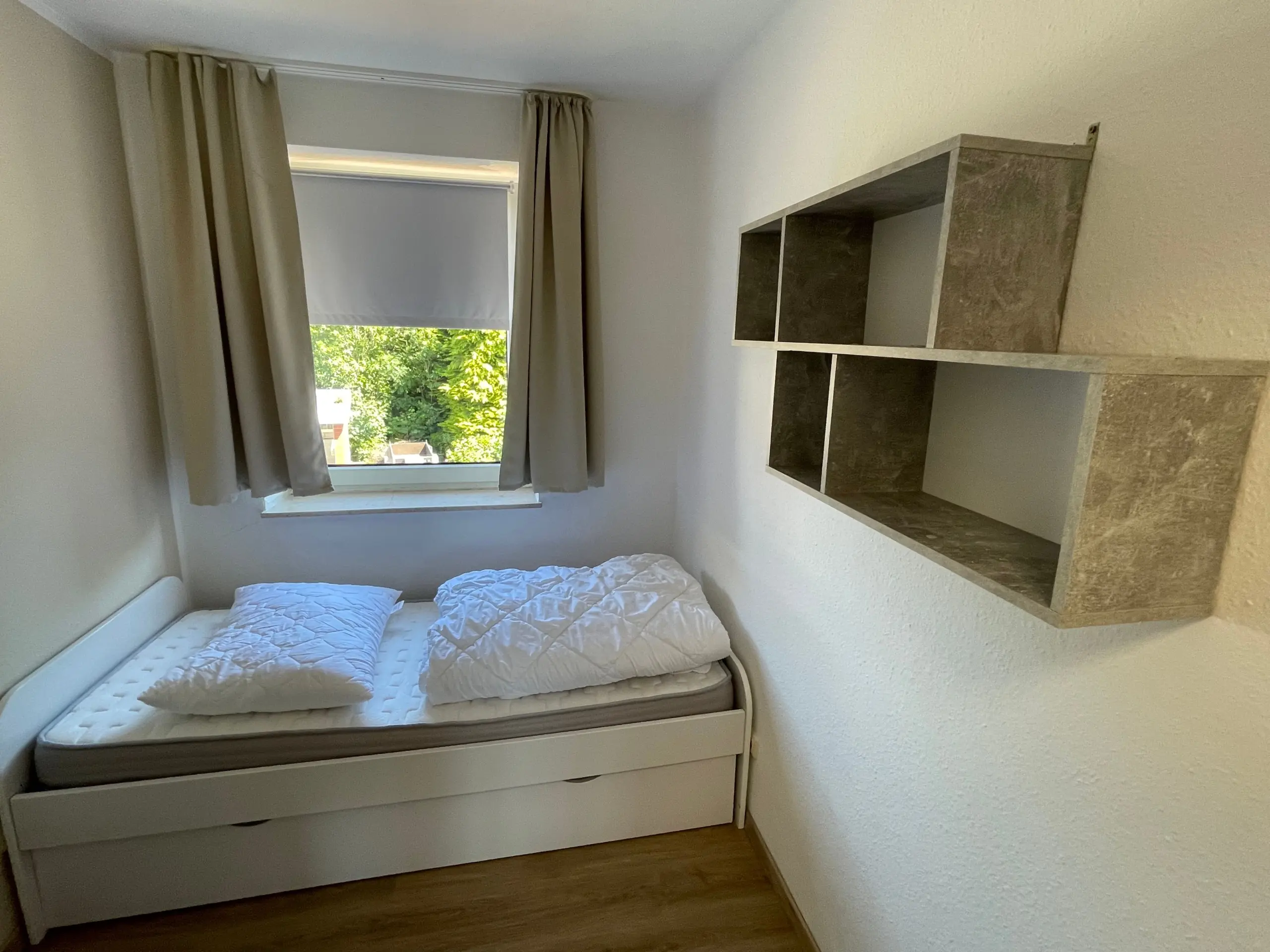 Small bedroom with a low, white bed and stacked white bedding, set against a light wall beside a window with beige curtains opening to a green outdoor view.