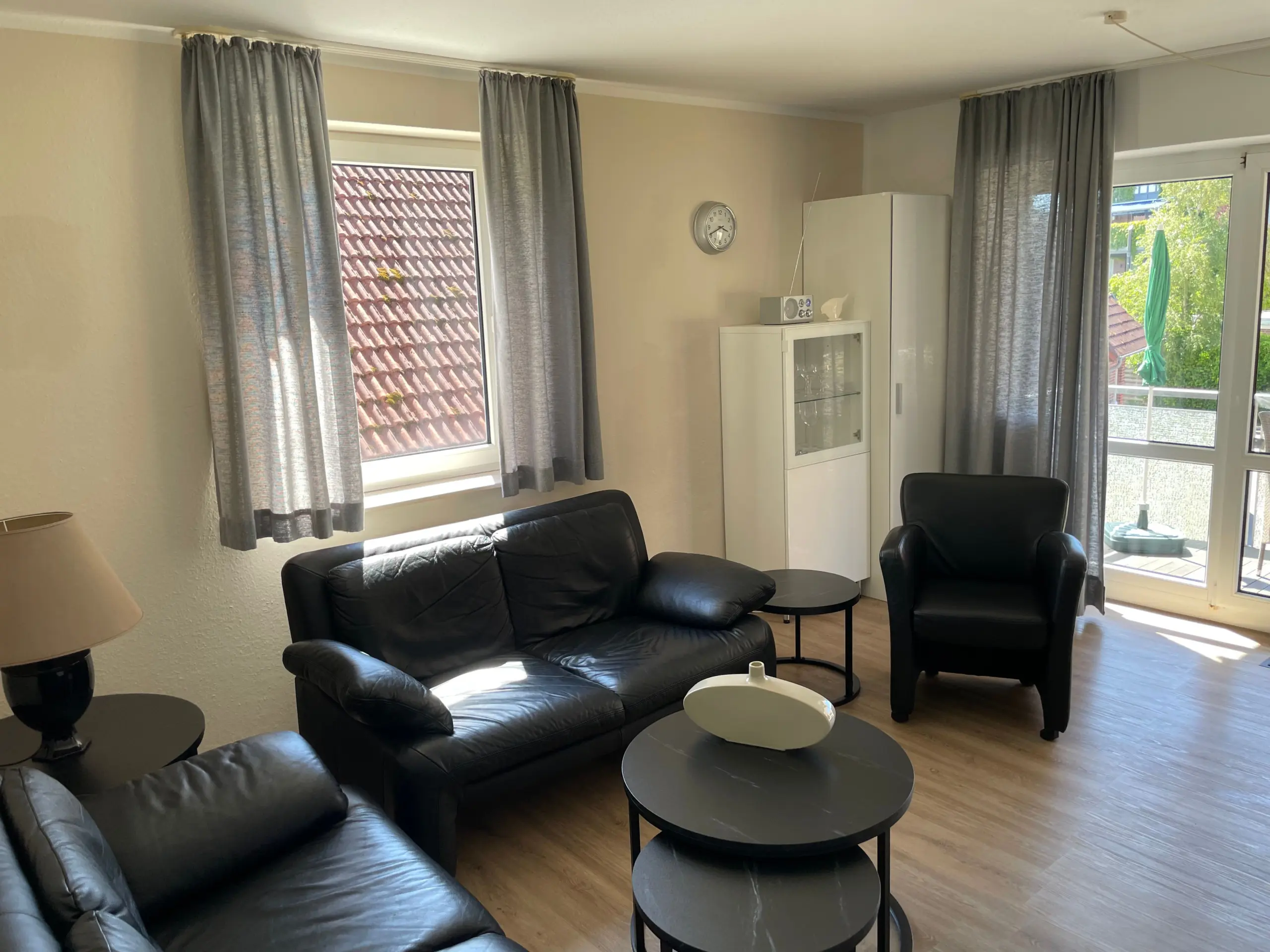 Modern living room with black leather sofa and chairs, two round black coffee tables, gray curtains, and a white cabinet near a window.