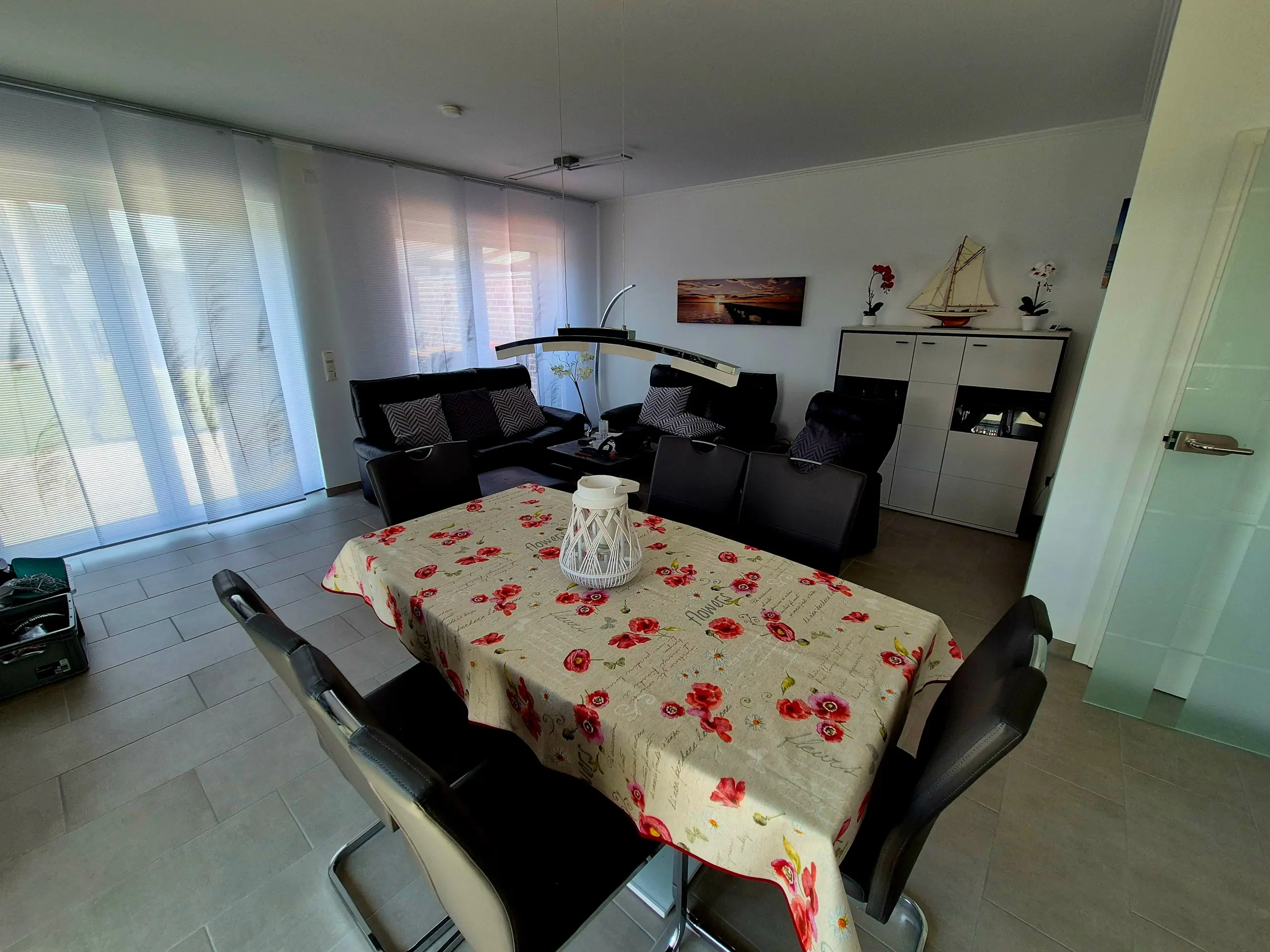 Dining area with a floral tablecloth and black chairs around a long table; large windows with blinds on the left.