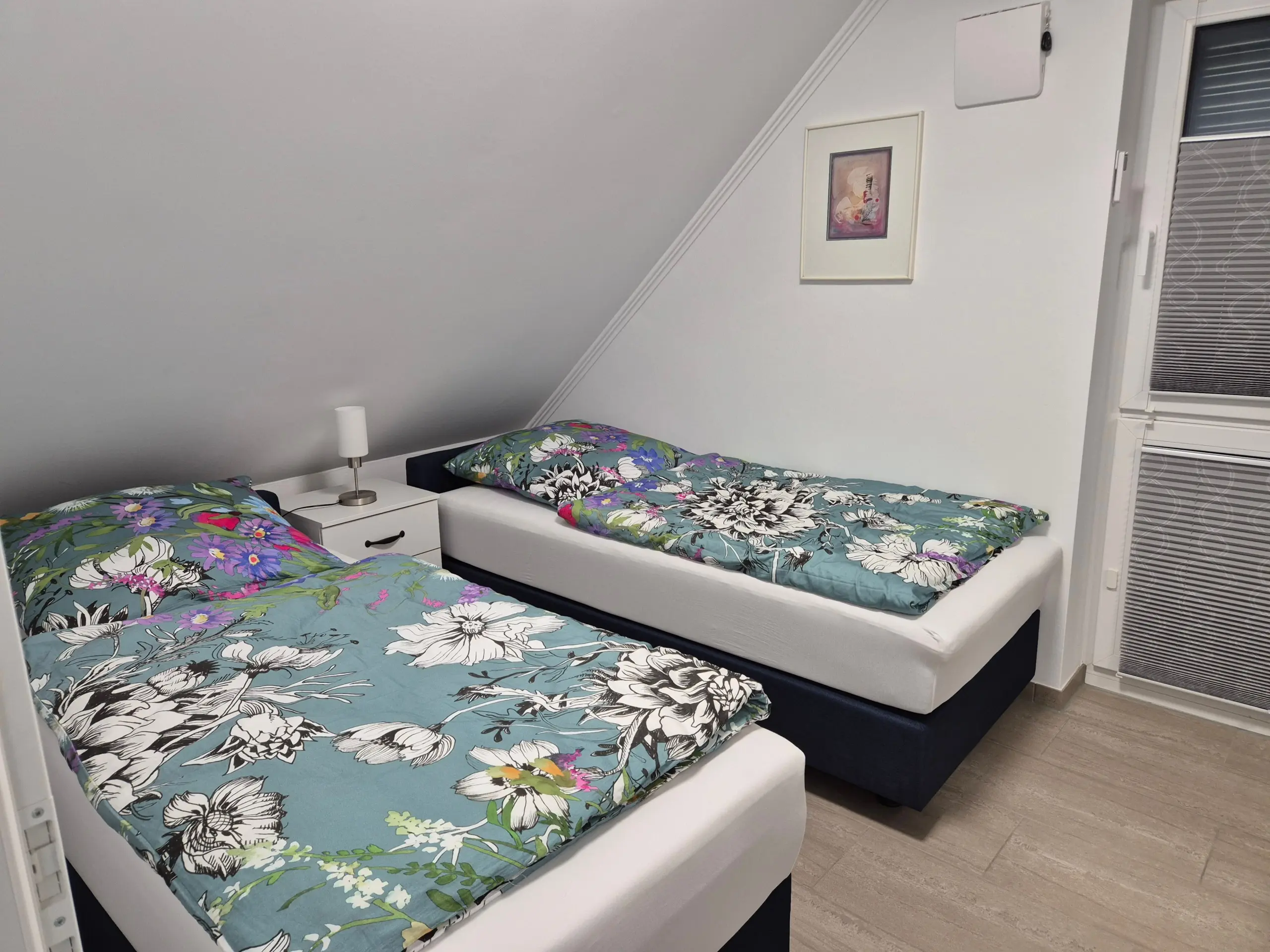 Twin beds with teal floral bedding and a white nightstand with lamp between them in a bright bedroom with a slanted wall and framed art above.