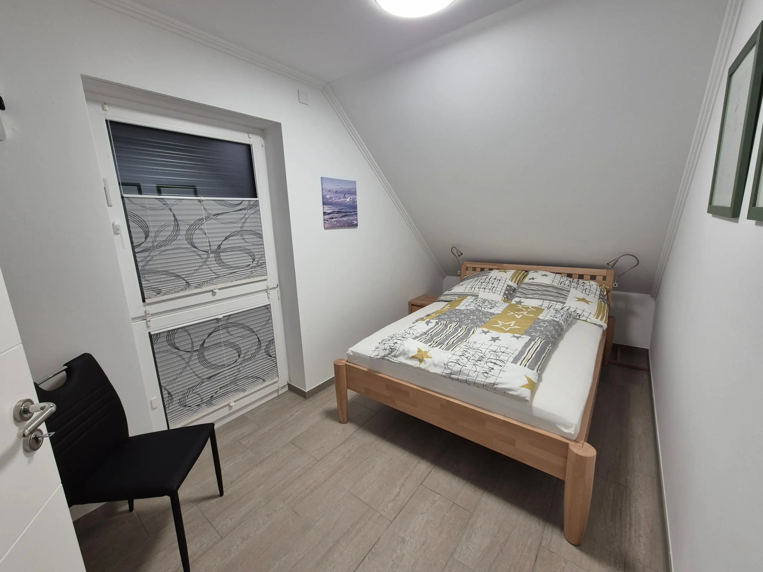 Small bedroom with a single wooden bed, two bedside tables, wall art, and a chair near the door under a sloped ceiling.