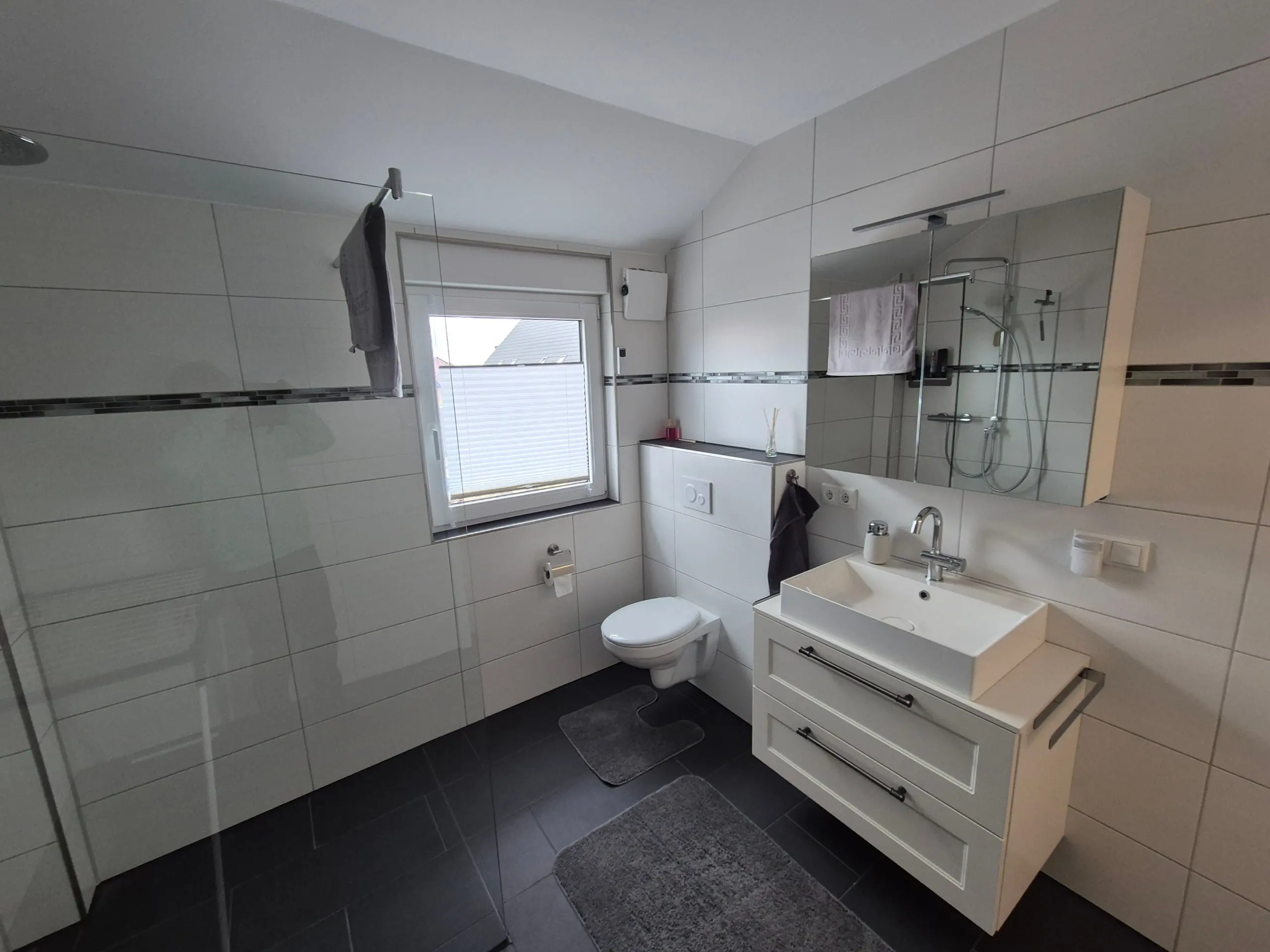 Modern white bathroom with wall-mounted vanity, large mirror, toilet, and glass shower; gray floor mats nearby on dark tile floor.