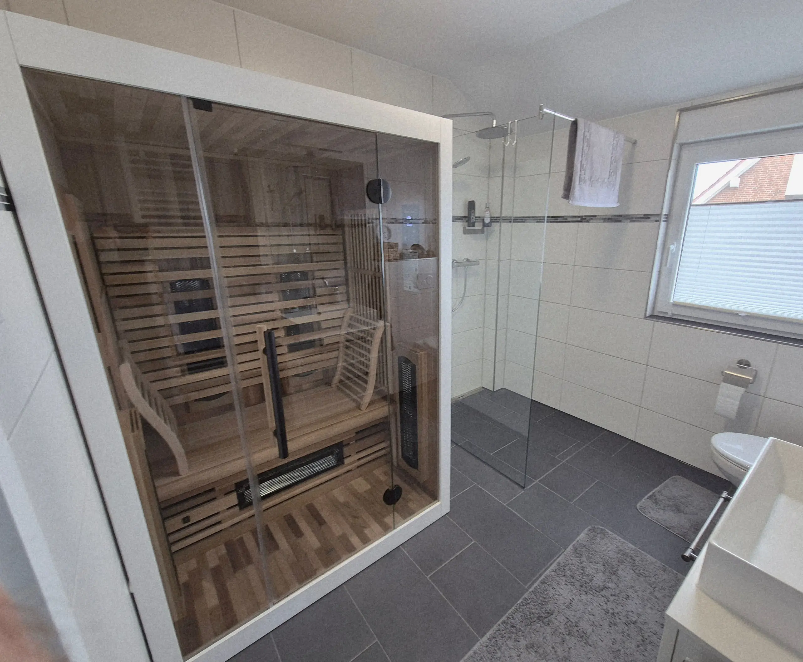 Glass-enclosed wooden sauna in a modern bathroom, with benches inside and a towel hanging near a windowed wall on the right.