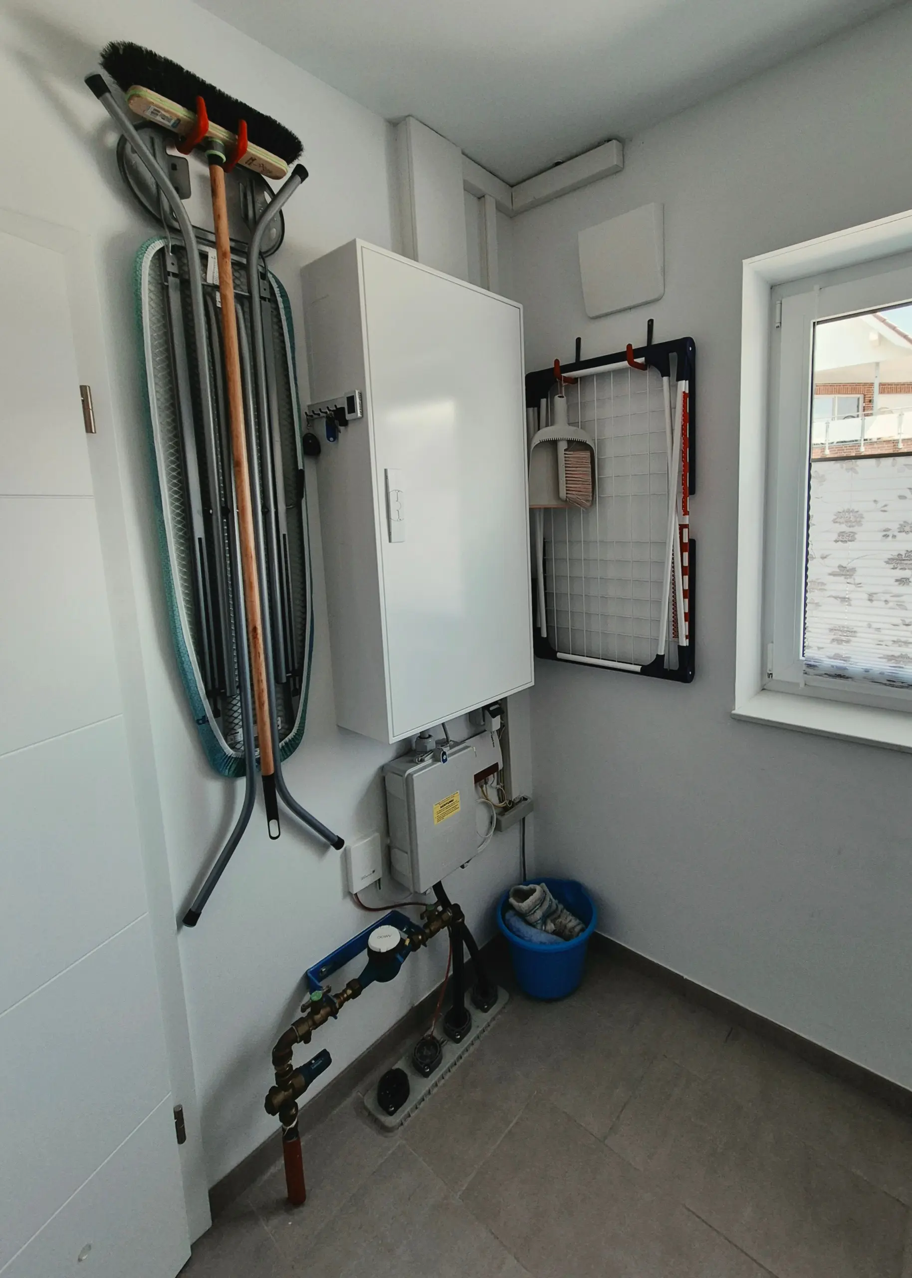 Utility room with cleaning tools stored on a wall rack (brooms, mop), a white cabinet, exposed pipes, and a window.