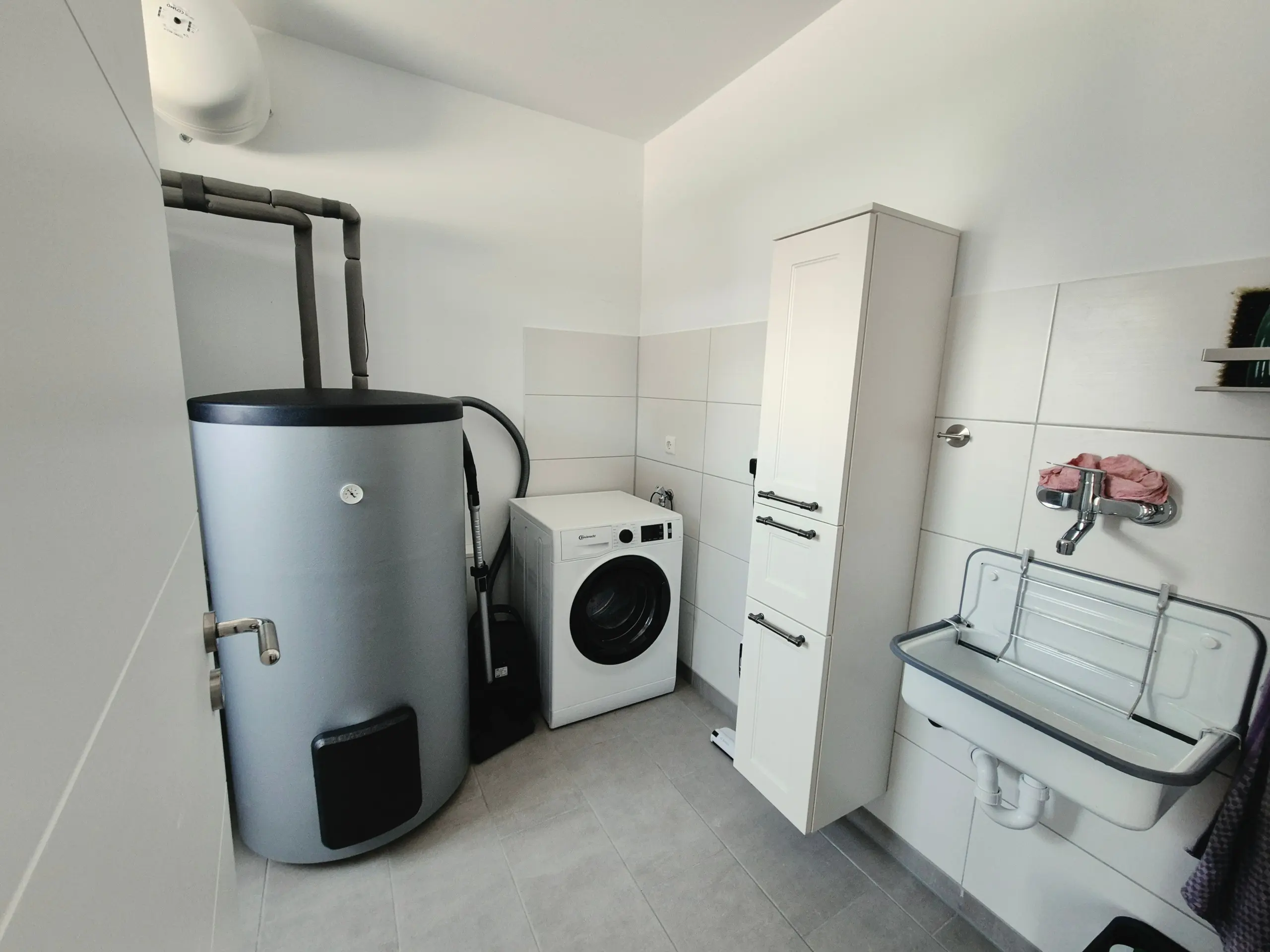 Narrow utility room with a tall white water heater, exposed gray pipes, a front-loading washing machine, and a white cabinet with sink nearby.