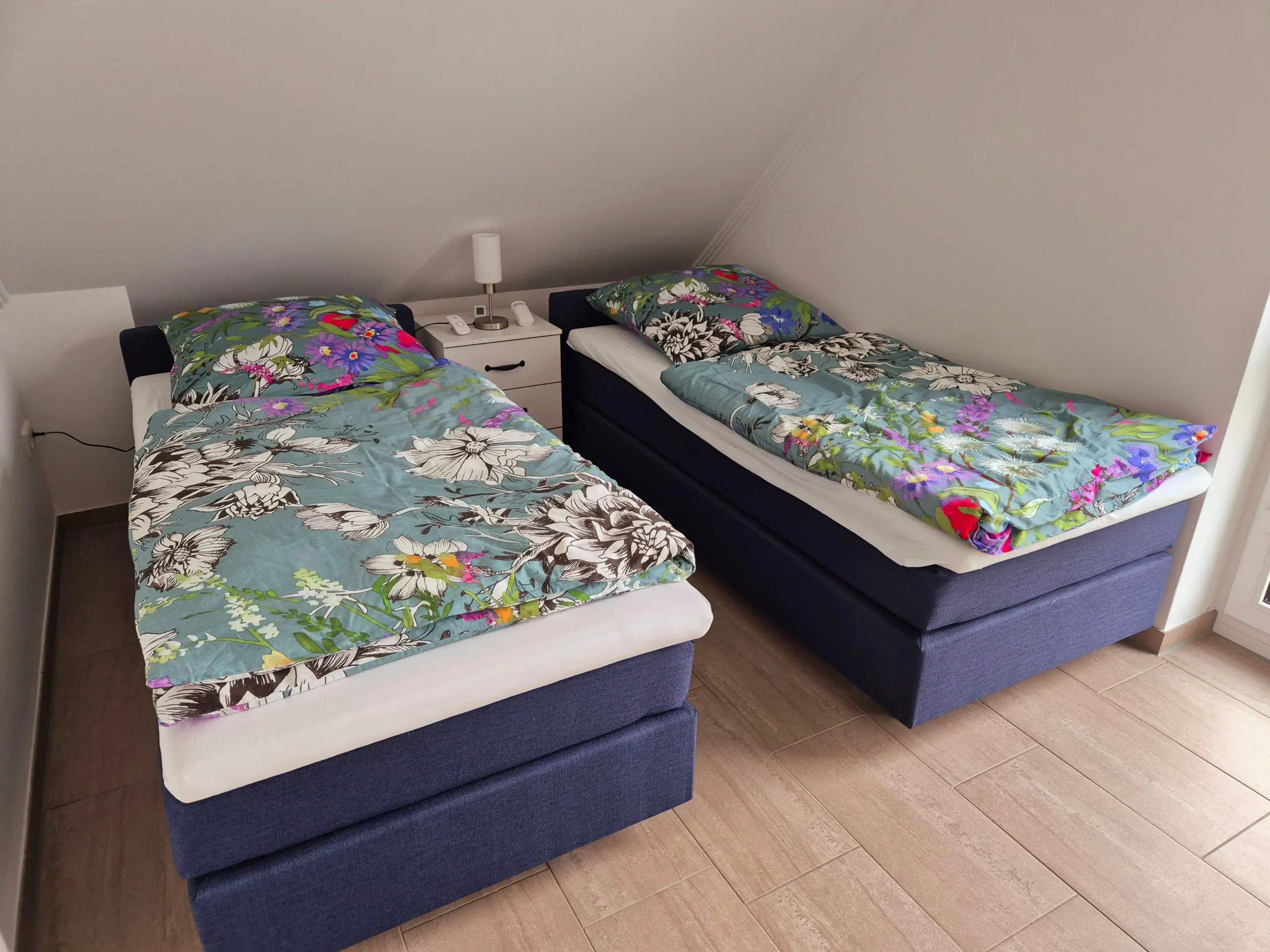 Two single beds with bright floral blue duvets and a white nightstand between them under a sloped ceiling.