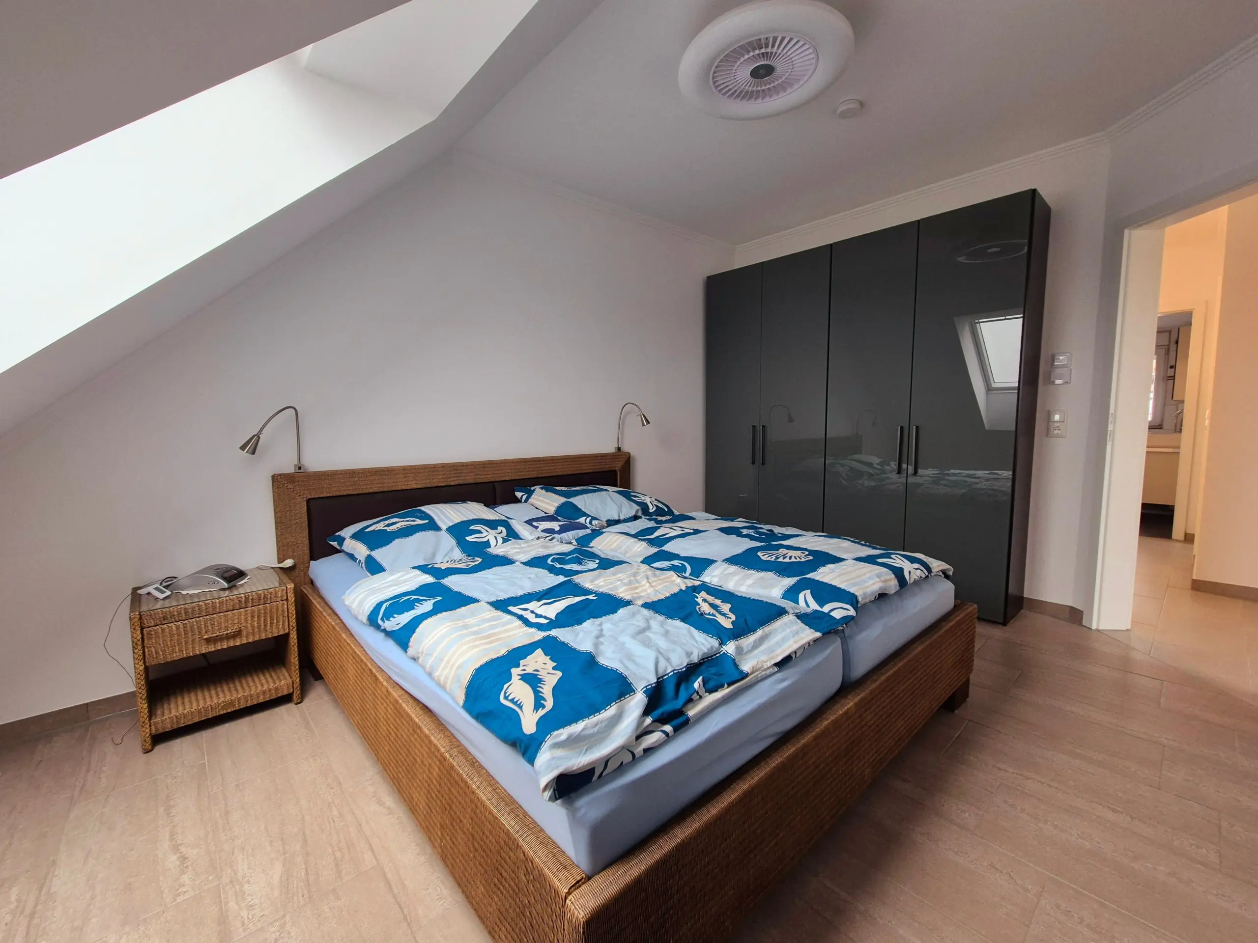 Bedroom with a wicker bed frame, blue seashell-pattern bedding, matching wicker nightstands, wall-mounted lamps, and a large dark wardrobe along a sloped ceiling wall.
