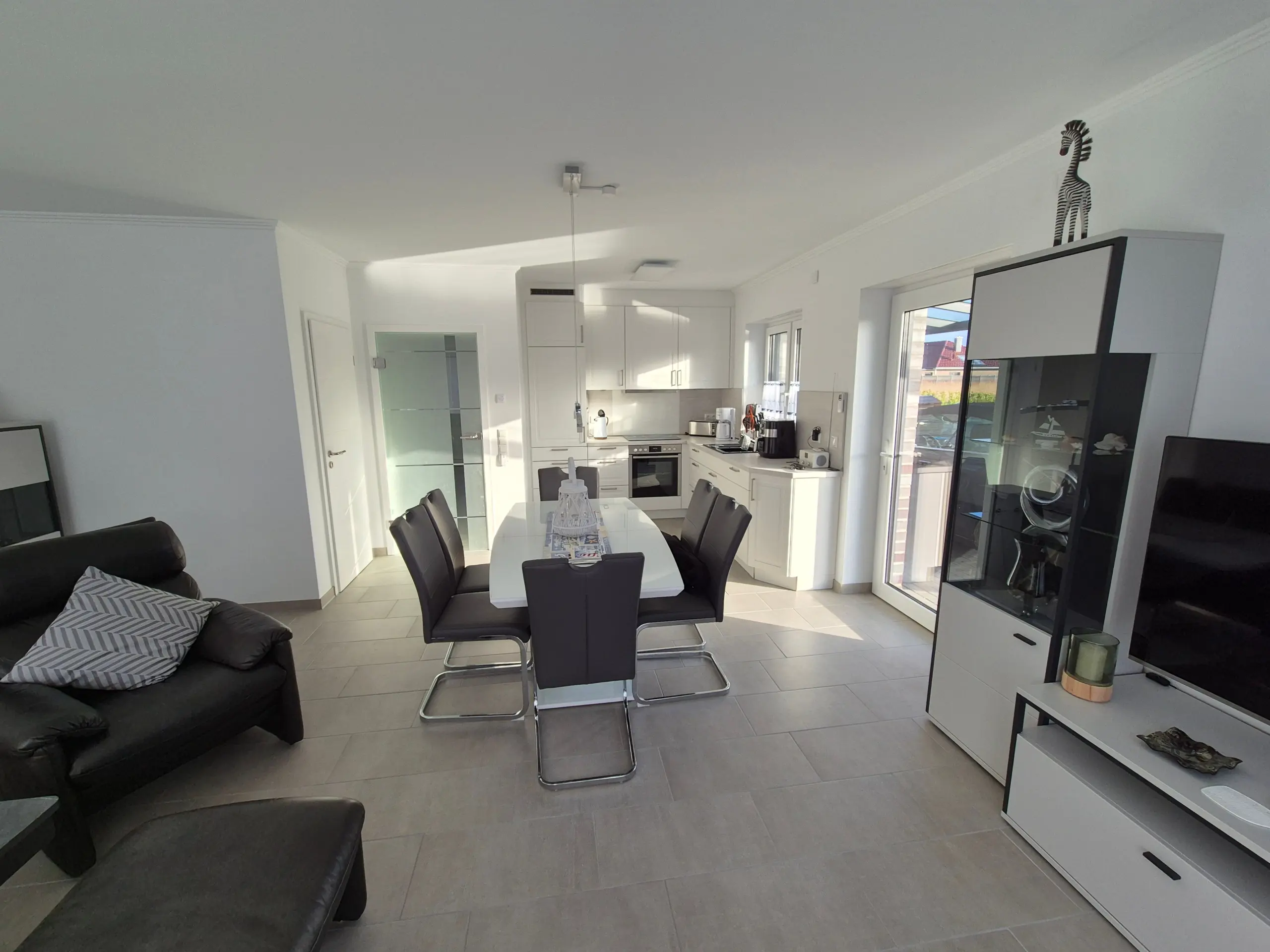 Bright open-plan kitchen and dining area with a long white table and black chairs, white cabinetry, and glass-front cabinets; zebra figurine atop a cabinet adds decor.