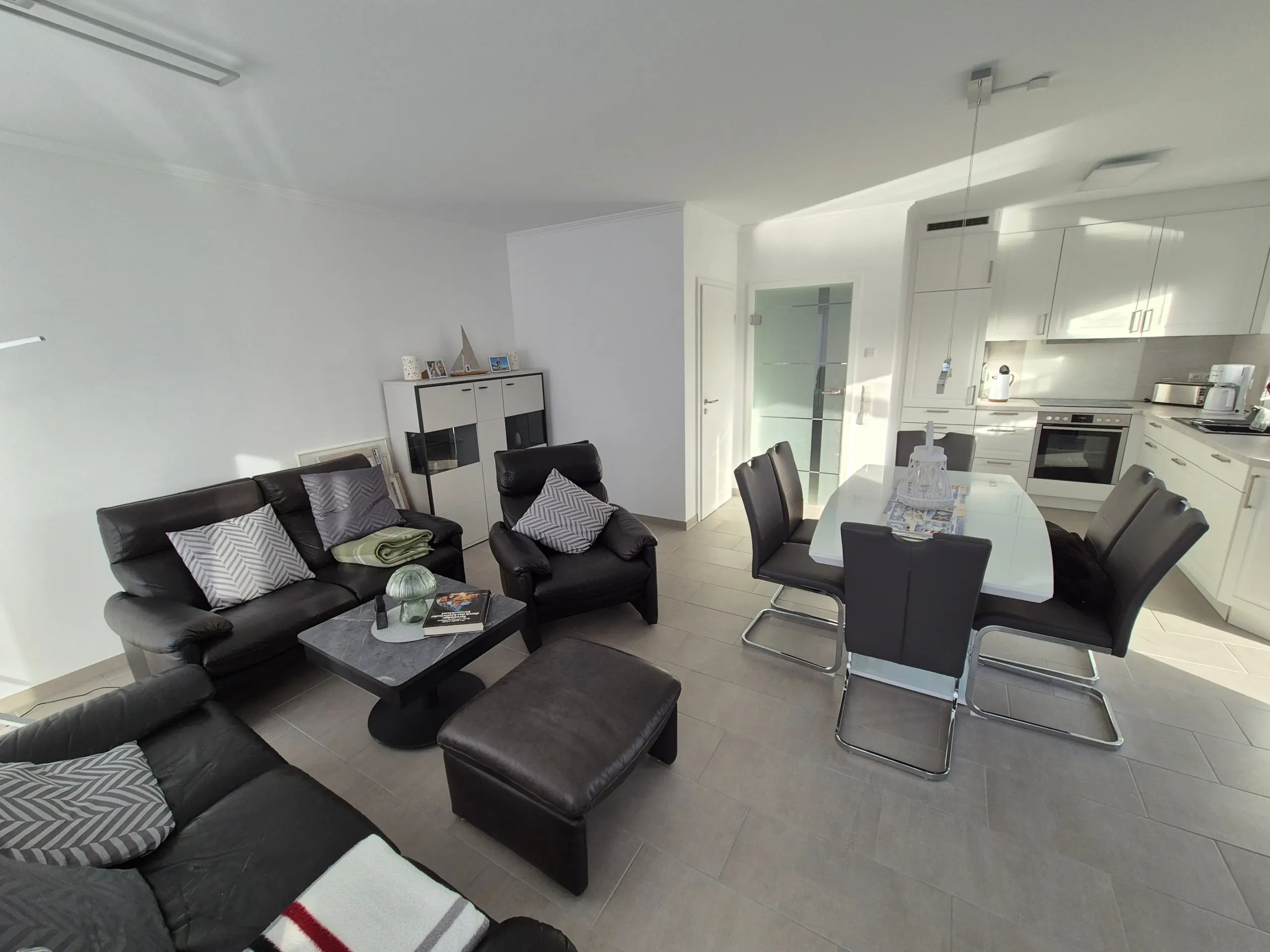 Open-plan living area with black leather sofas, grey cushions, and a white kitchen along the right wall, in a bright, modern home.