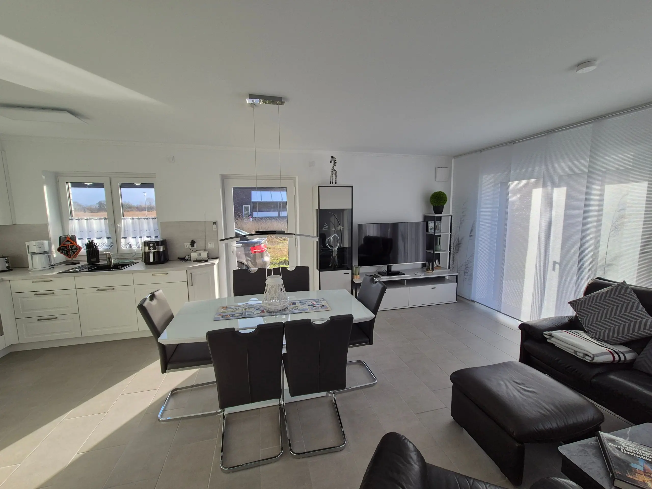 Bright open-plan kitchen and living room with white cabinets, glass dining table, black chairs, TV unit, and a zebra statue on a cabinet.