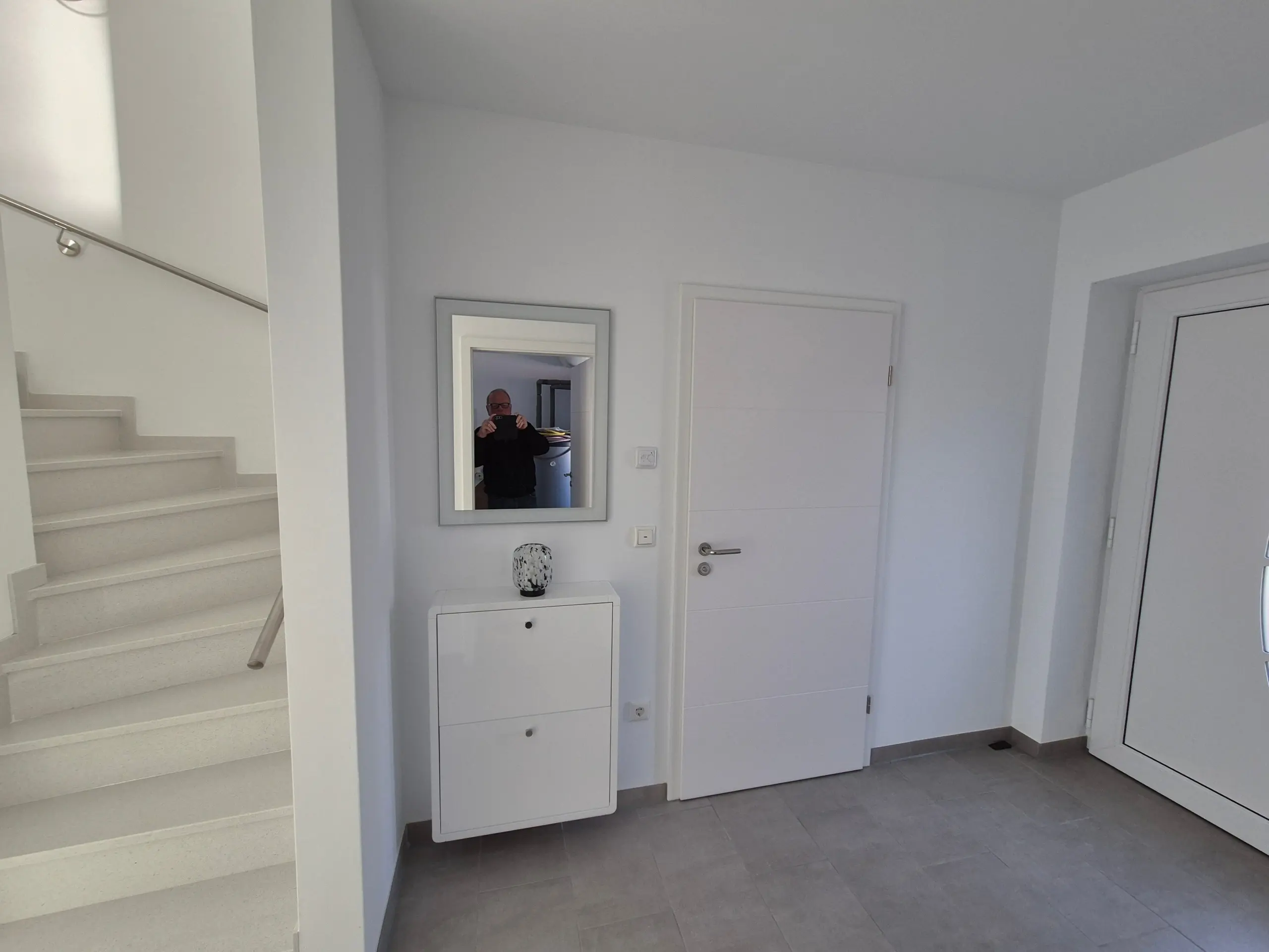 White, modern hallway with stairs on the left, a small cabinet with a vase, and a mirror reflecting a person taking a photo.