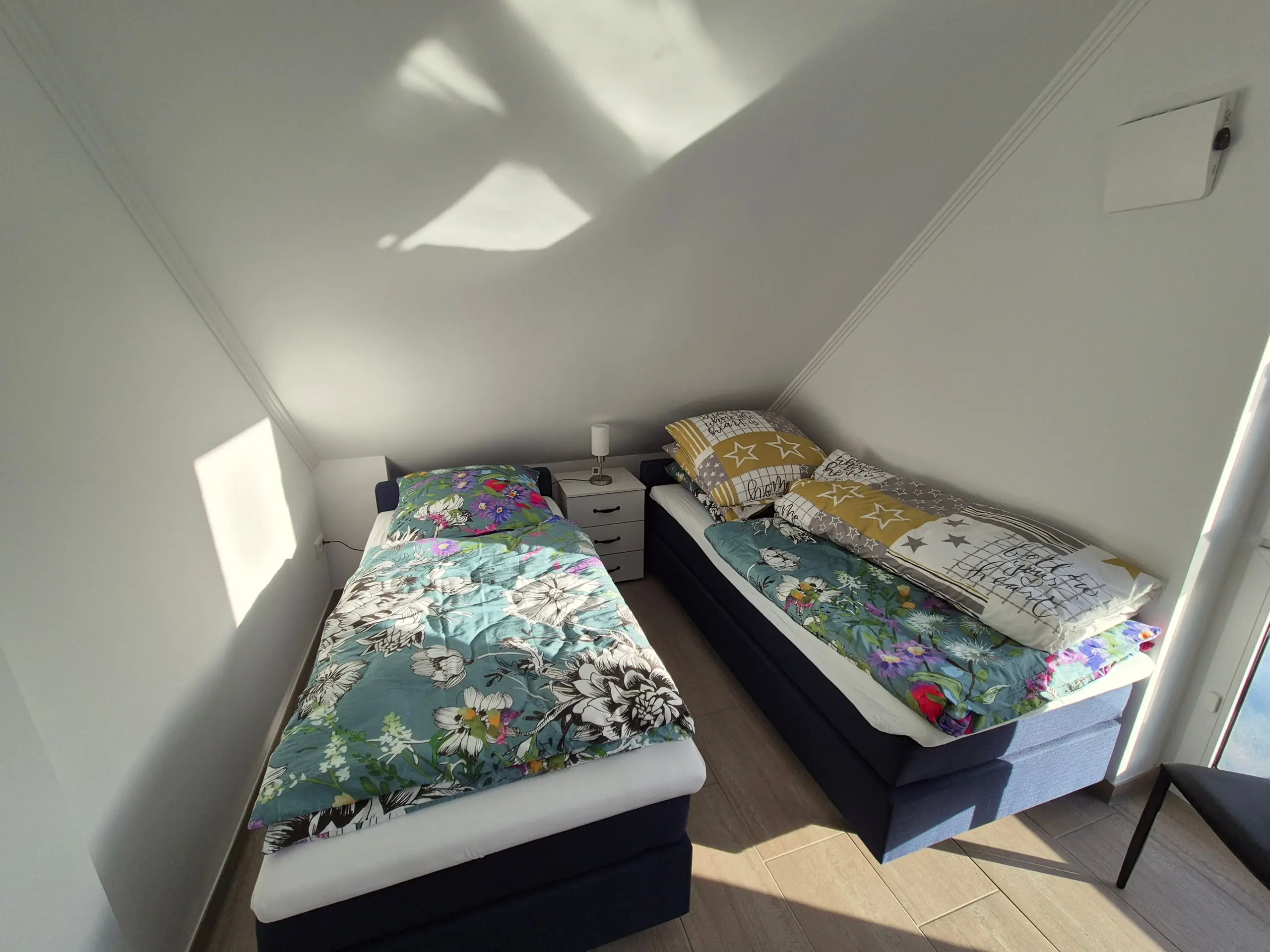 Two twin beds with colorful floral quilts sit in a small attic bedroom, separated by a white nightstand and lamp under a sloped ceiling.