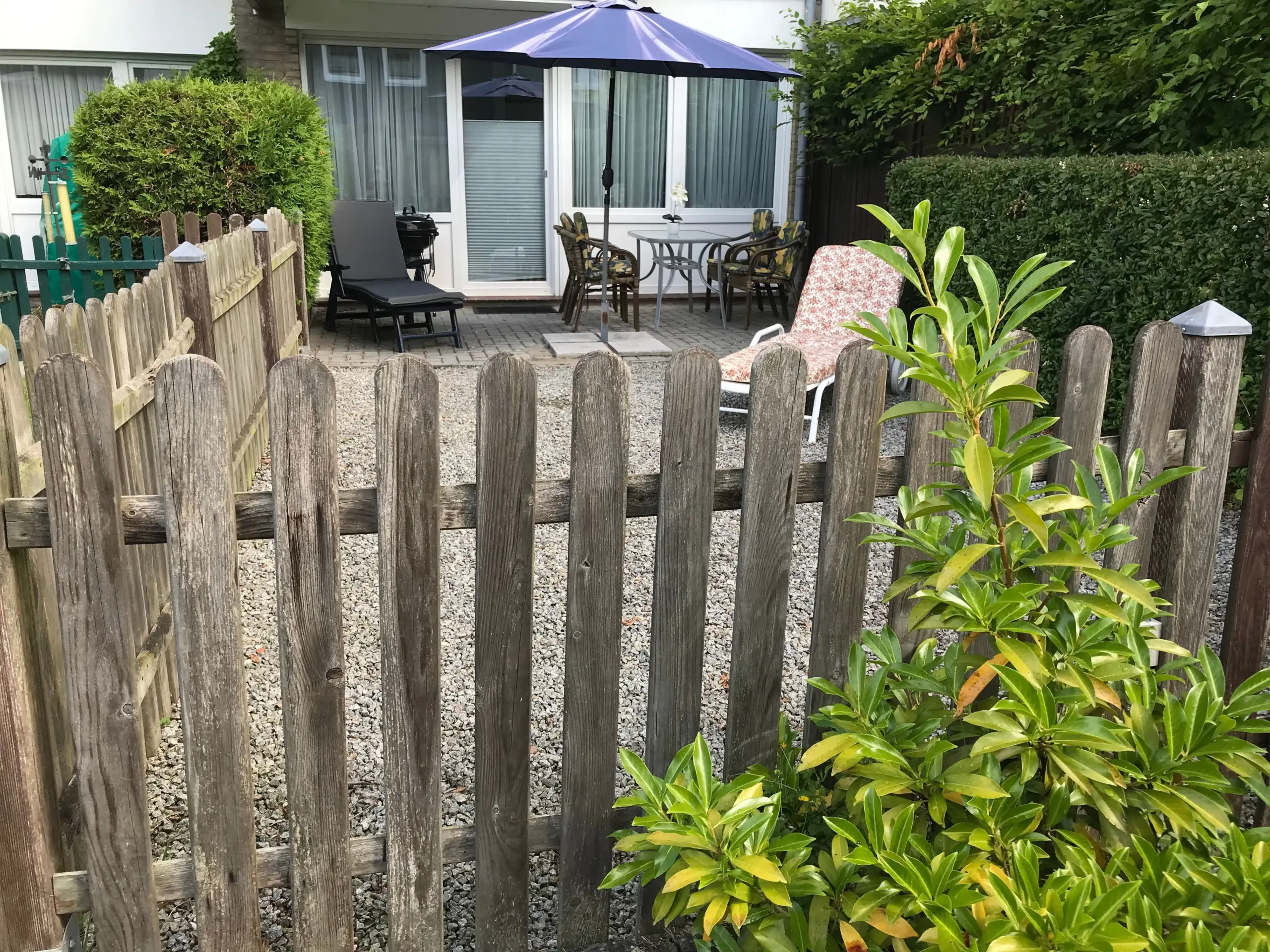 Backyard patio with a blue umbrella over a round table and four chairs, a lounge chair nearby, and sliding glass doors in the background.