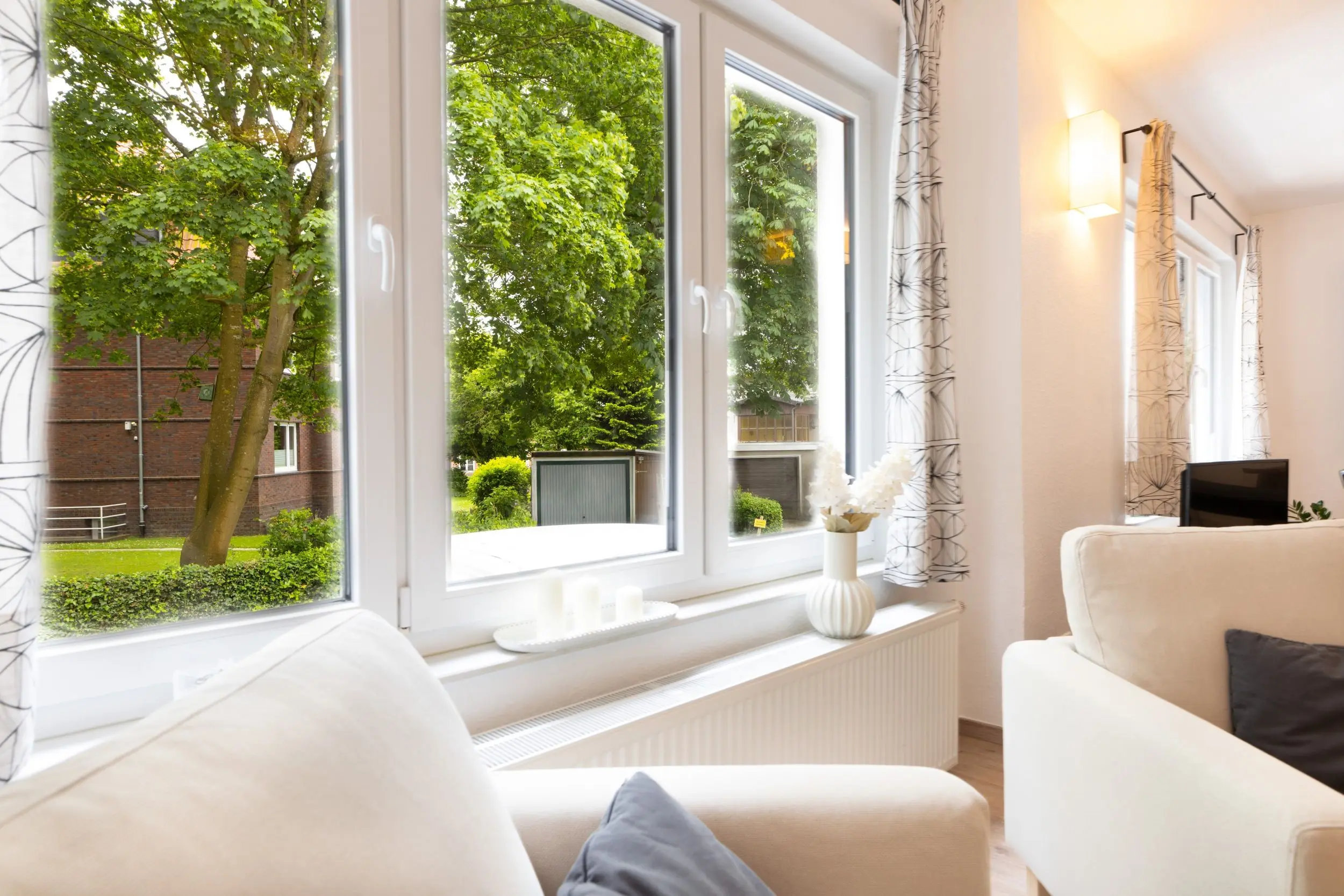 Bright living room with large white-framed windows showing green trees outside, and white furniture inside.