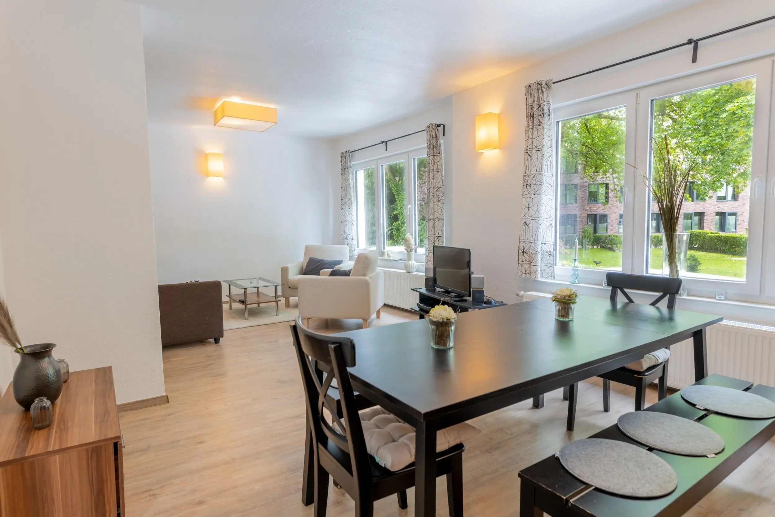 Bright open-plan living and dining area with a black dining table, grey cushioned stools, and large windows overlooking greenery outside telegraphing natural light and a modern space.