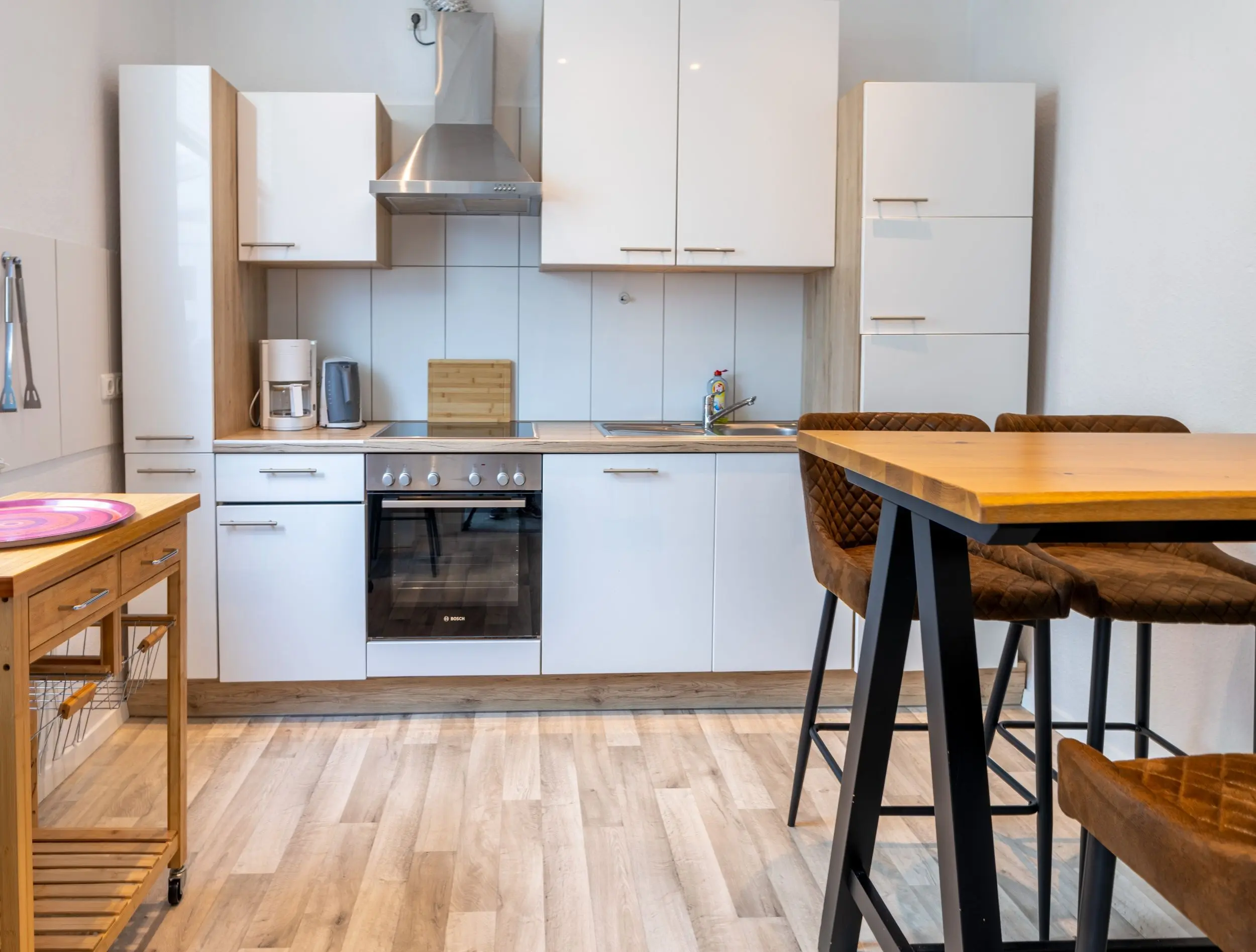 Modern kitchen with white cabinets, stainless steel range hood, built-in oven, and a wooden dining table with black metal stools.
