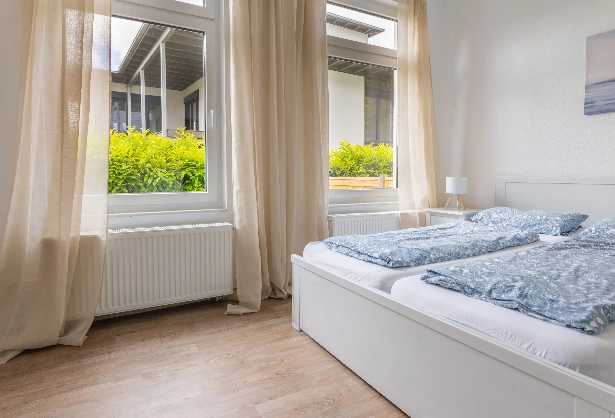 Bright bedroom with two white twin beds, blue patterned duvets, beige curtains, and a view of greenery outside the windows.