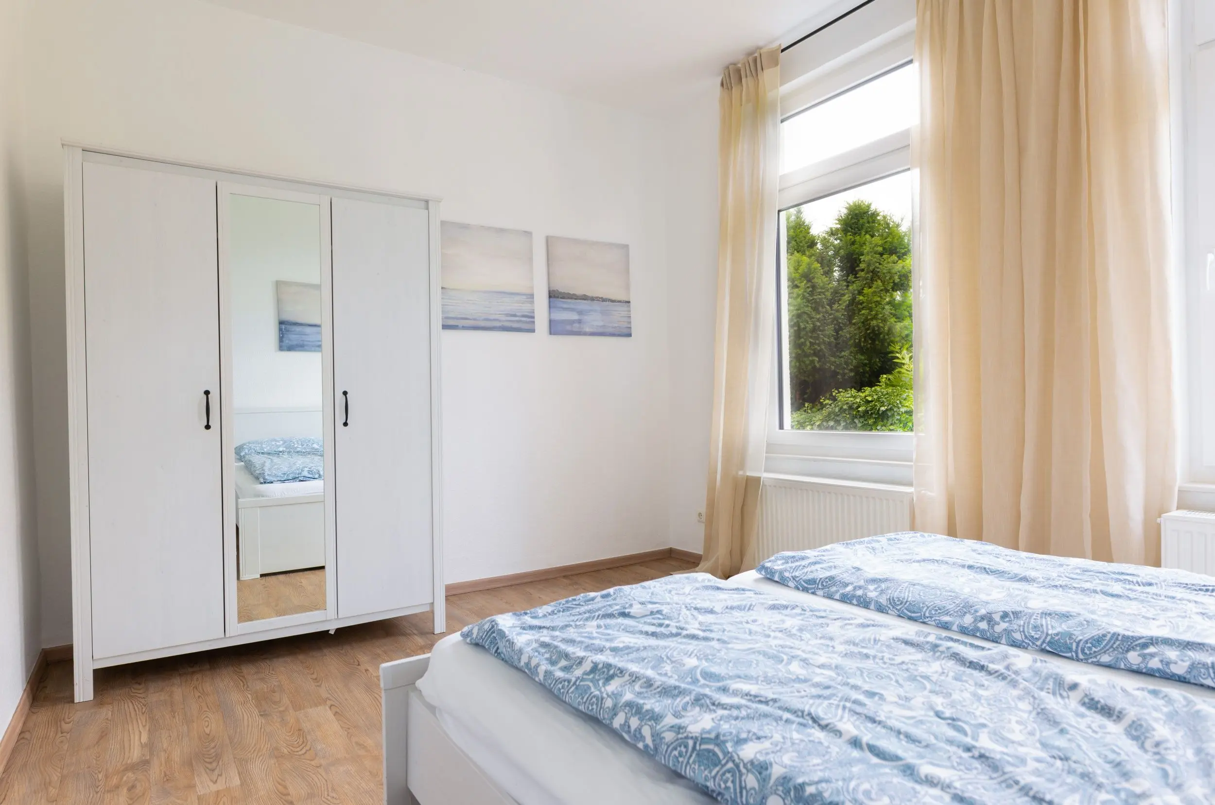 Bright bedroom with white wardrobe, mirrored door, two blue-patterned beds, and a large window with beige curtains letting in daylight.