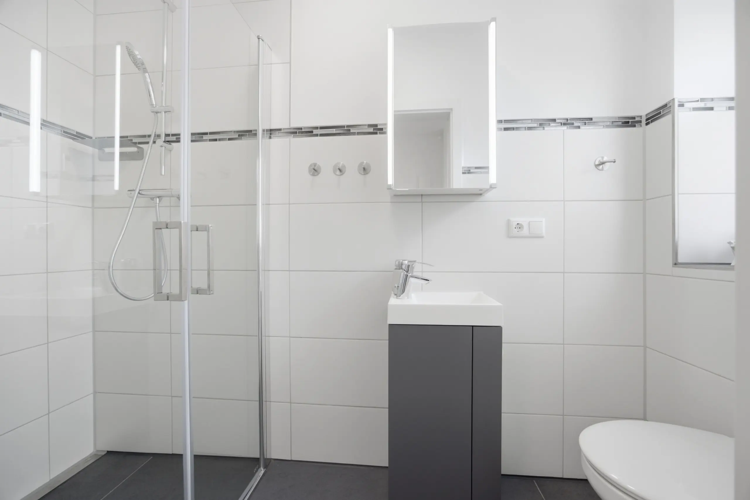 Modern white bathroom with glass shower enclosure, wall mirror cabinet, and square sink on a dark vanity; minimal chrome fixtures.