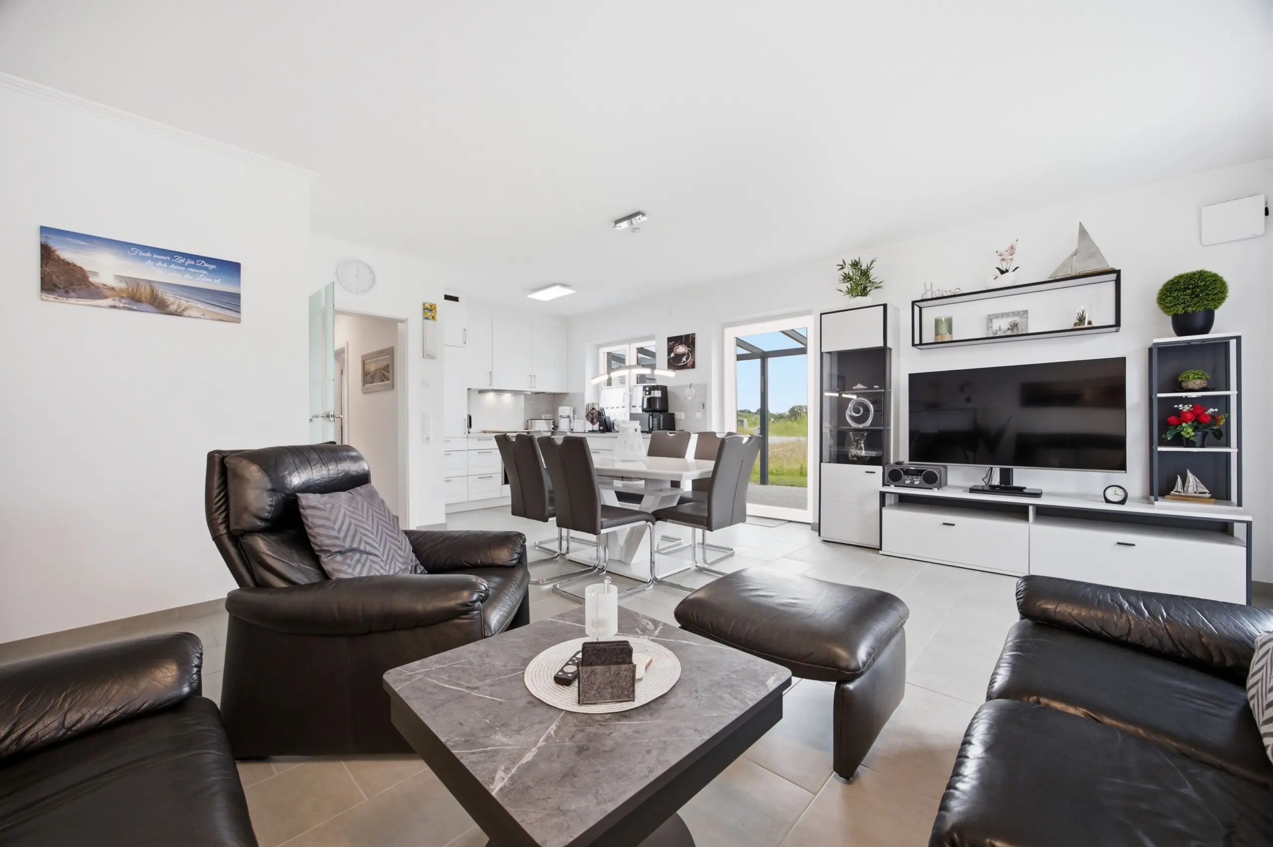 Open-plan living room with black leather sofas around a dark coffee table, white walls, and a wall-mounted TV; kitchen and dining area visible beyond.