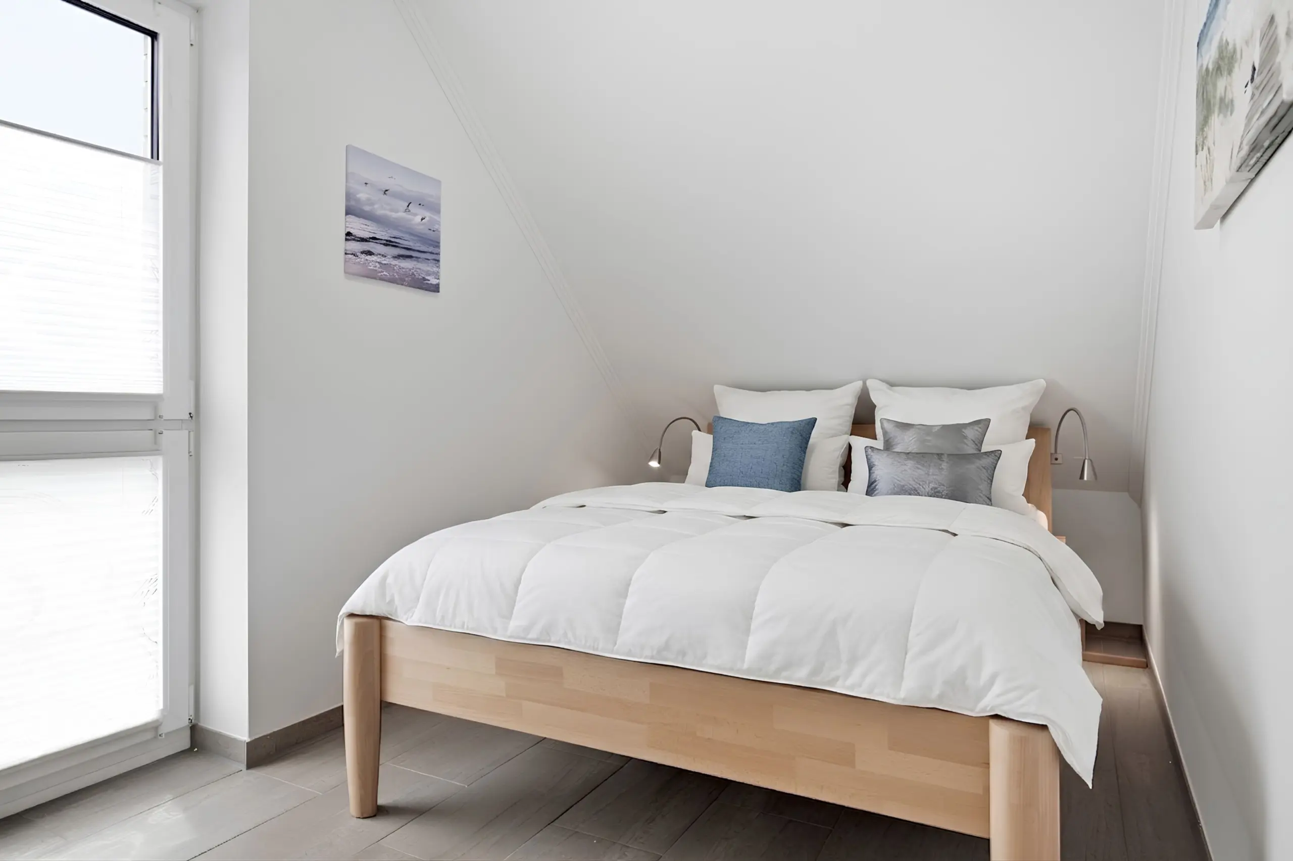 Bedroom with a wooden platform bed, white bedding, and blue/gray pillows against a light wall with wall art on the left and a window with blinds to the side.