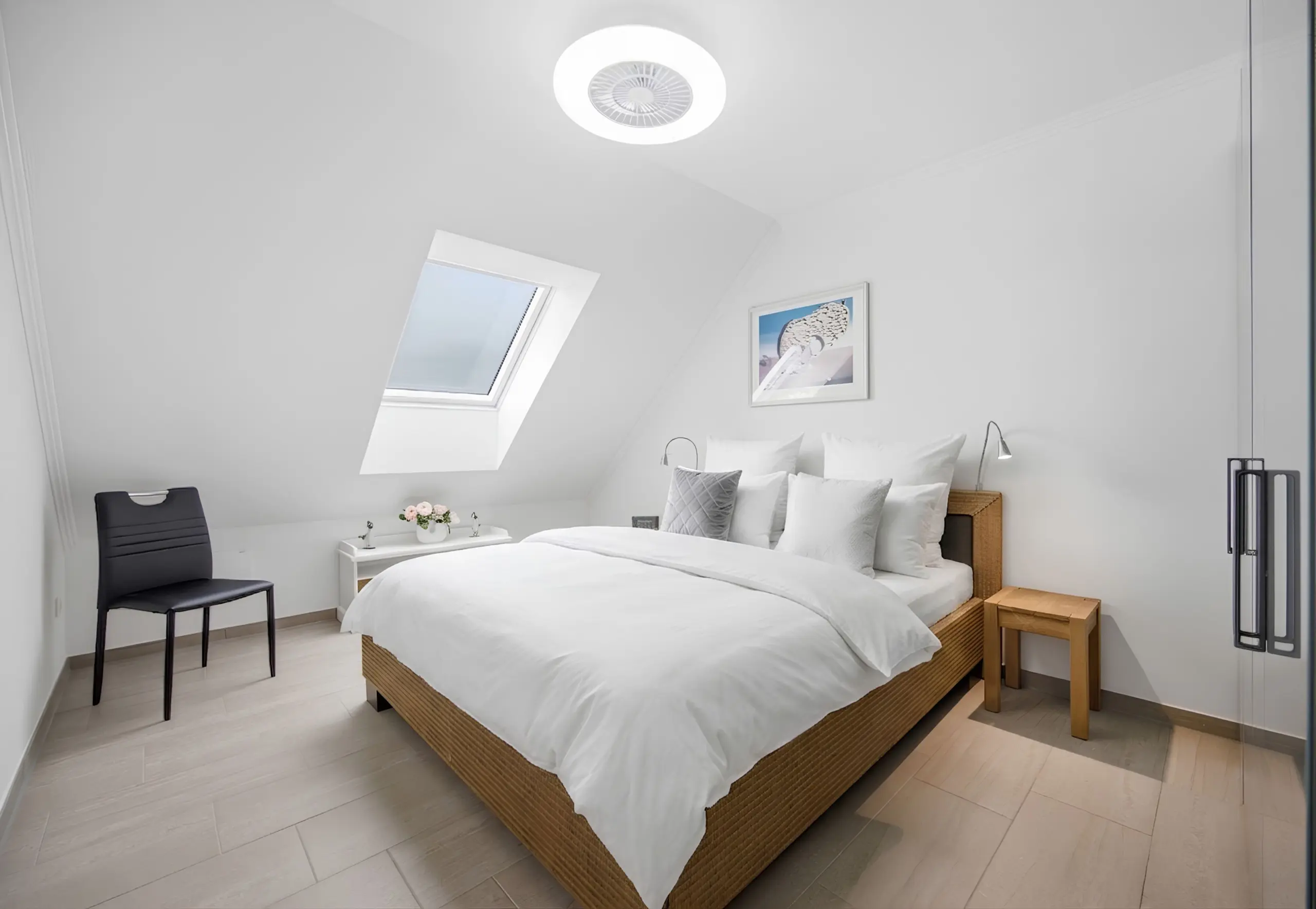 Bright attic bedroom with white bedding, wicker bed frame, skylight, and minimalist décor including a black chair.