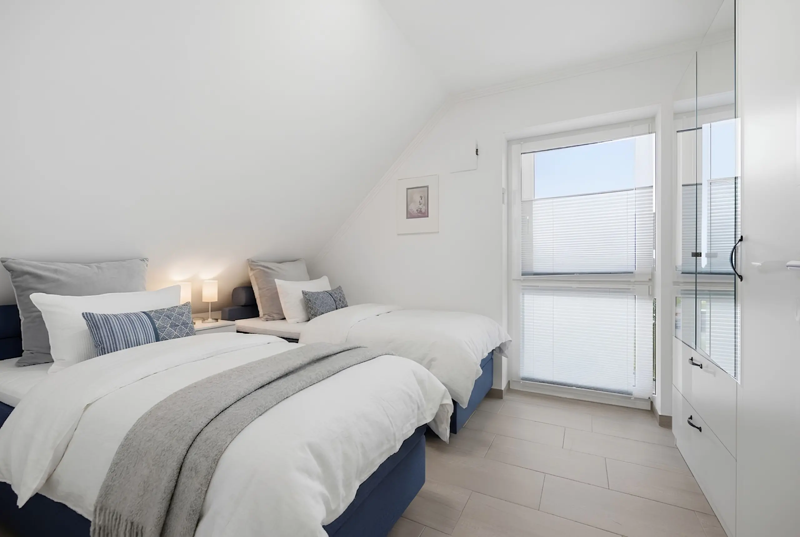 Bright white bedroom with two neatly made single beds, gray throws, and patterned pillows; large windowed wall on the right.