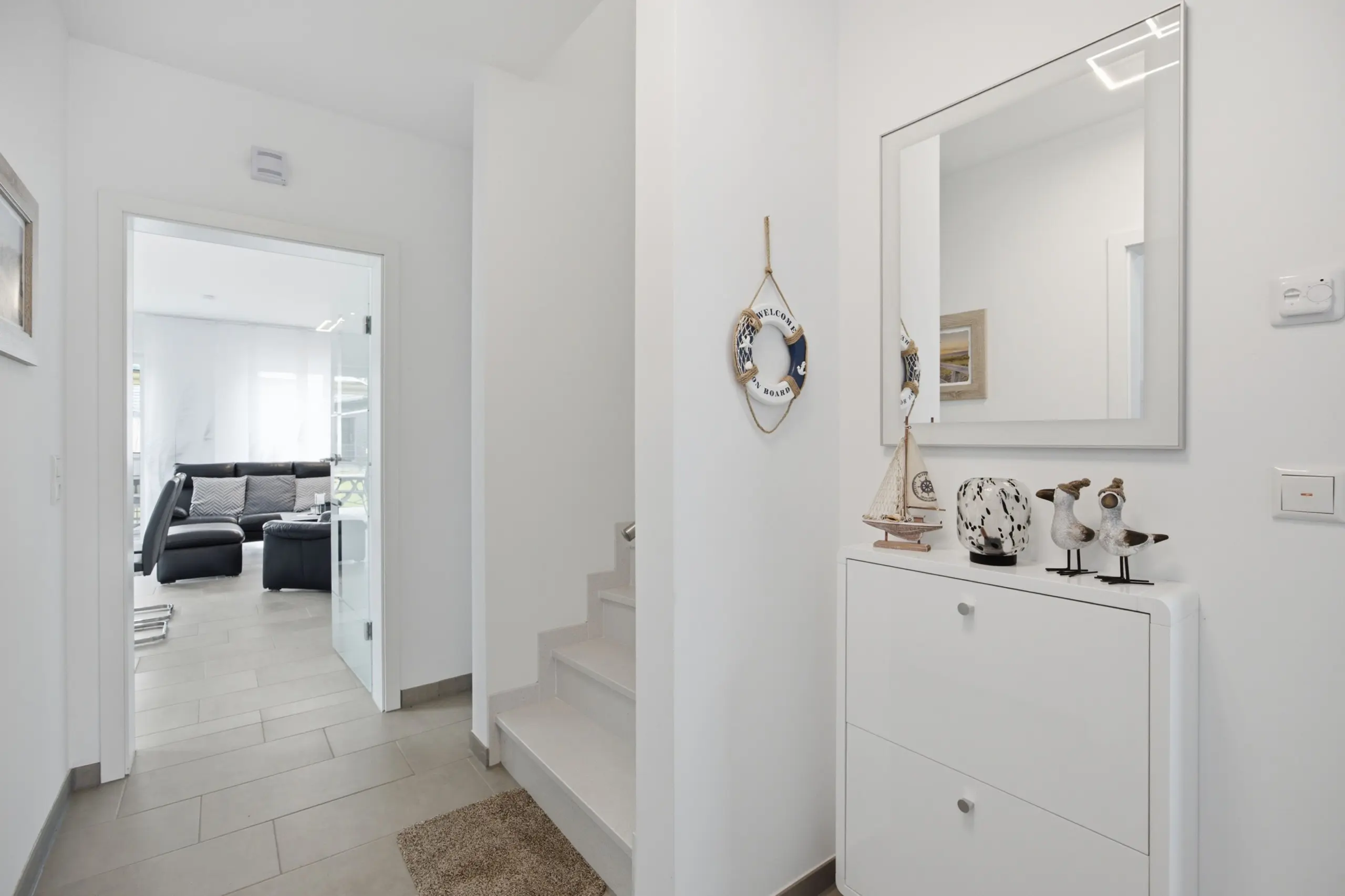 Light, white hallway with stairs on the right and a mirrored wall; decor includes sea-themed ornaments on a white cabinet and a lifebuoy hanging on the wall; living room visible through open doorway with a black sofa.