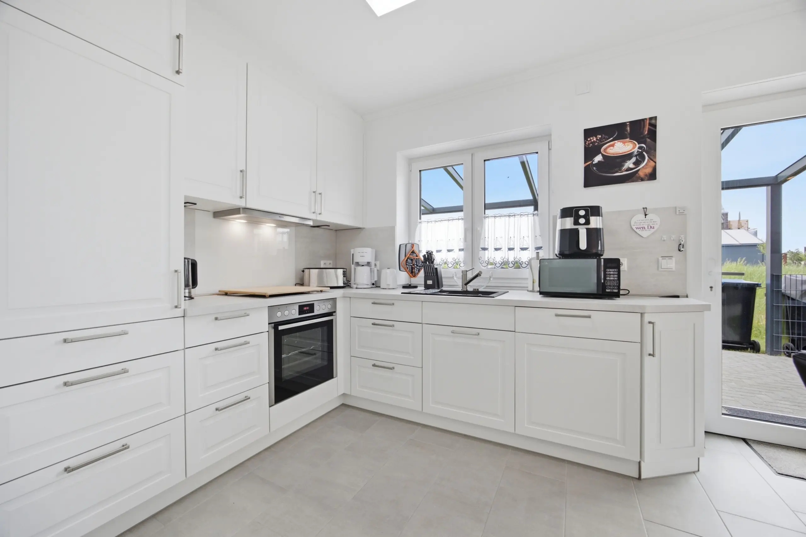 Bright white L-shaped kitchen with tall cabinets, stainless handles, and built-in oven under counter. A window and door lead outside; counter clutter minimal with small appliances.