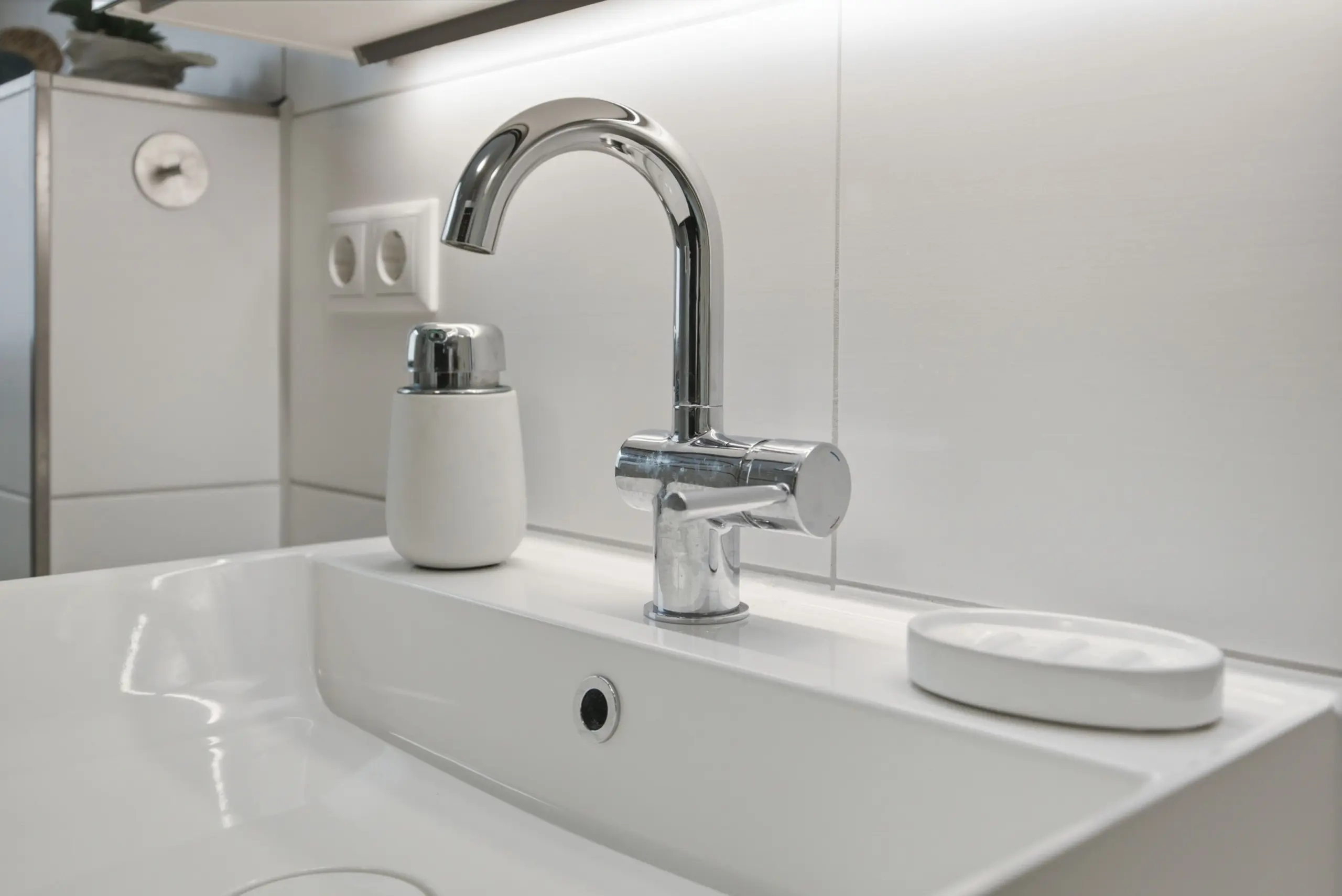 Chrome curved faucet over a white sink with a white soap dispenser on a glossy countertop in a bright modern bathroom surface, tiled wall in background.