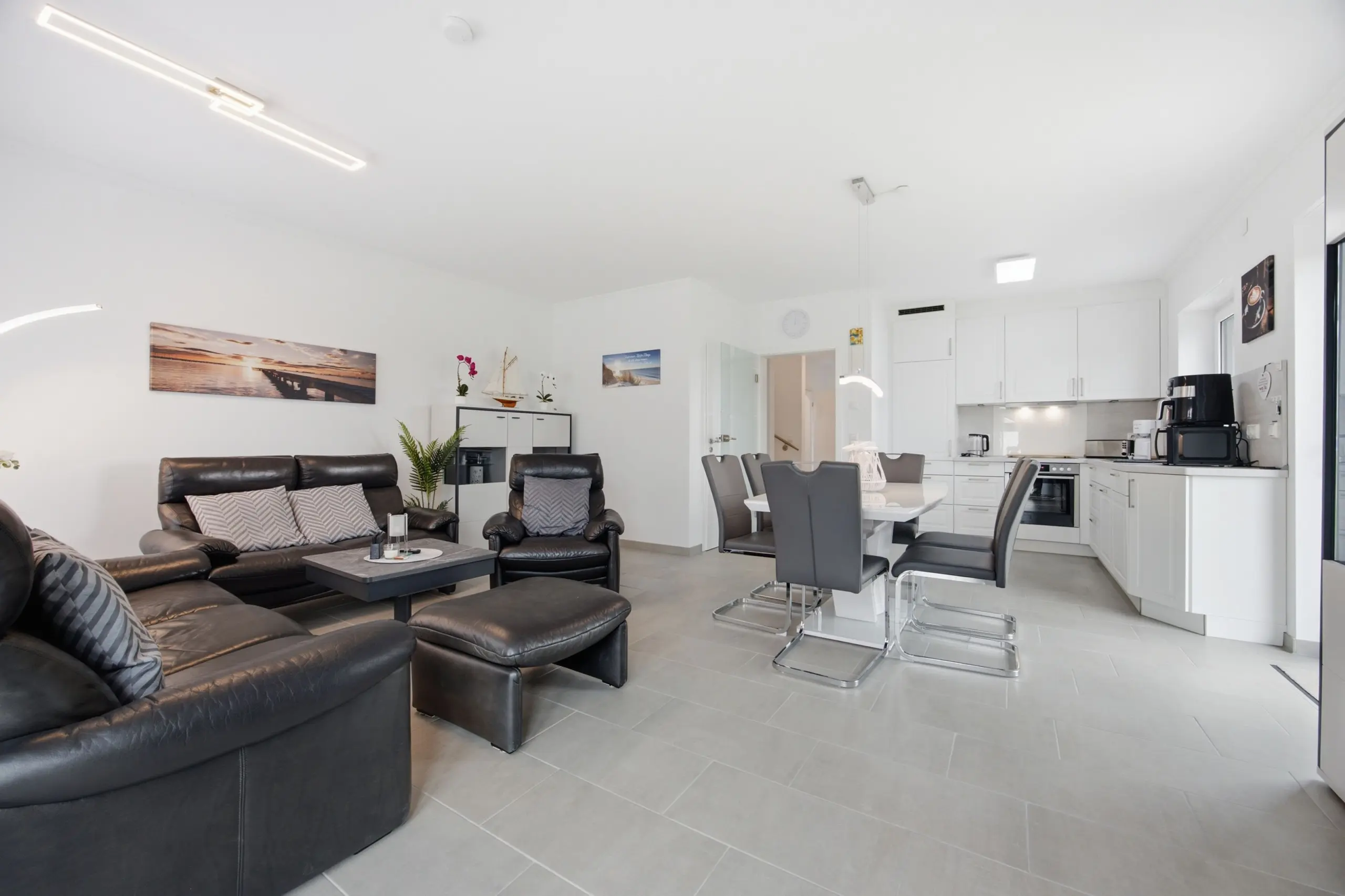 Open-concept living room with dark leather sofas, coffee table, and a white kitchen in the background.