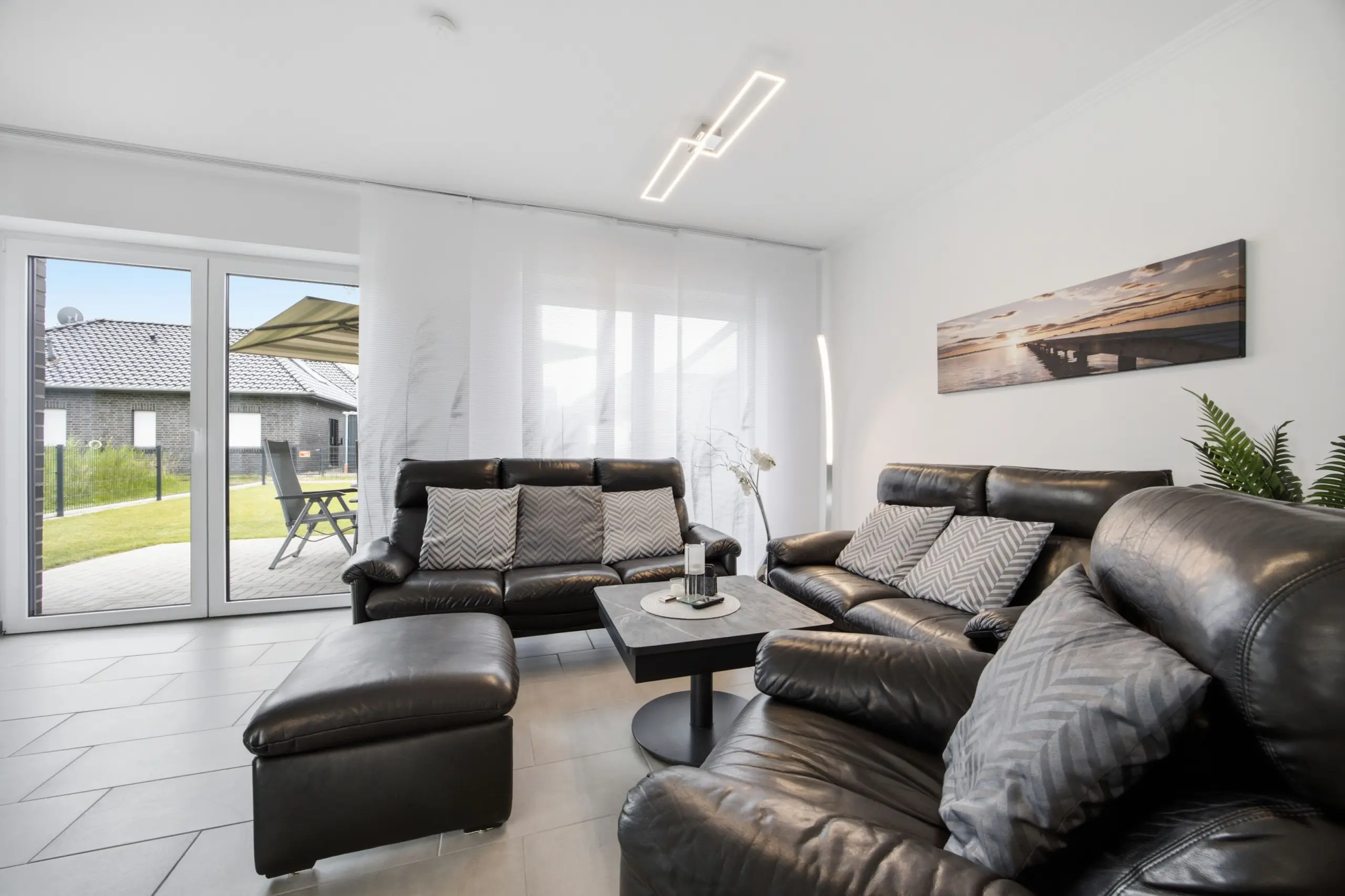 Living room with black leather sectional sofa, striped throw pillows, and a glass coffee table facing a large windowed wall. Outside, a patio with a table and umbrella is visible. A landscape photo hangs on the wall.