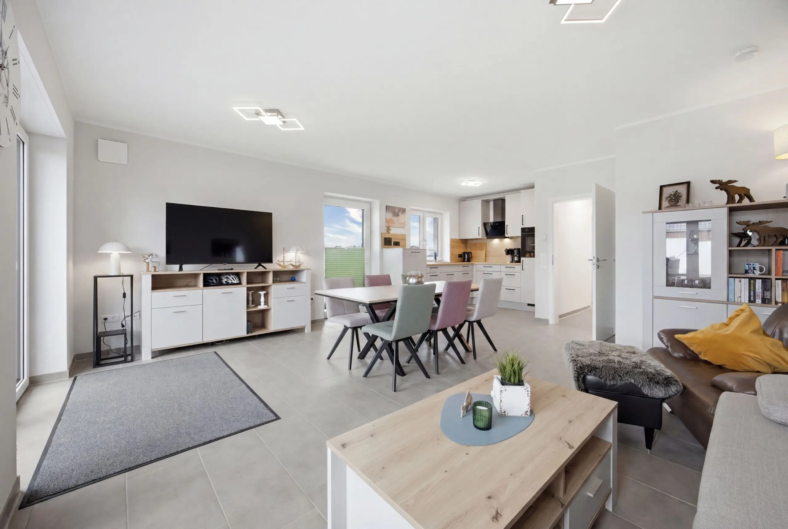 Open-plan living area with a wall-mounted TV, white cabinetry, and a dining table with pastel chairs under bright lights.