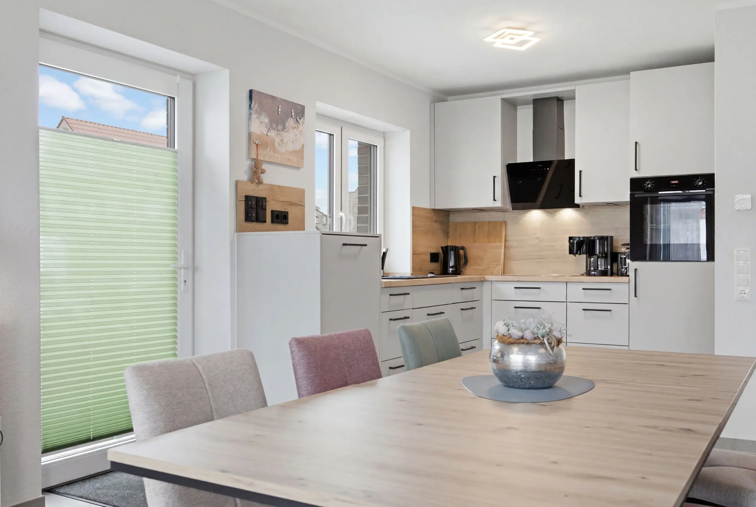 Bright modern kitchen with white cabinets, stainless steel oven, and a wooden dining table in the foreground.