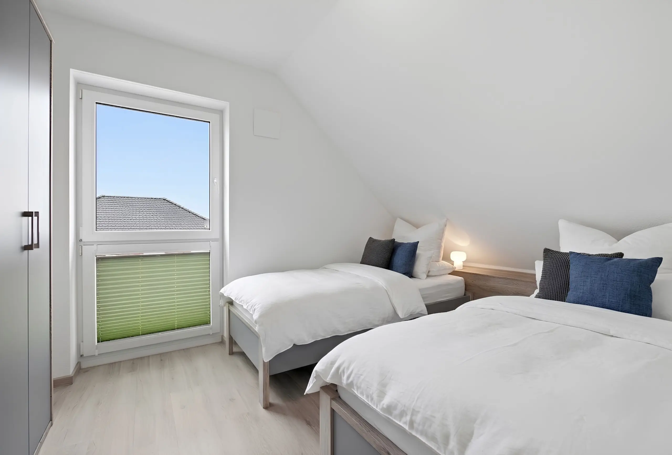 Bright modern bedroom with two single beds, white bedding, gray bed frames, and a window letting in daylight.